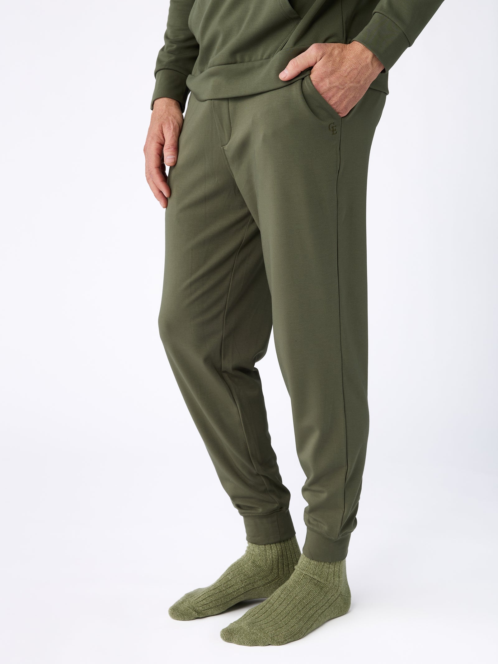 A person dressed completely in Cozy Earth's Men's Ultra-Soft Bamboo Pullover Crew in green stands against a white background. They have their hands in the pockets of their olive green pants and are wearing matching green socks. 