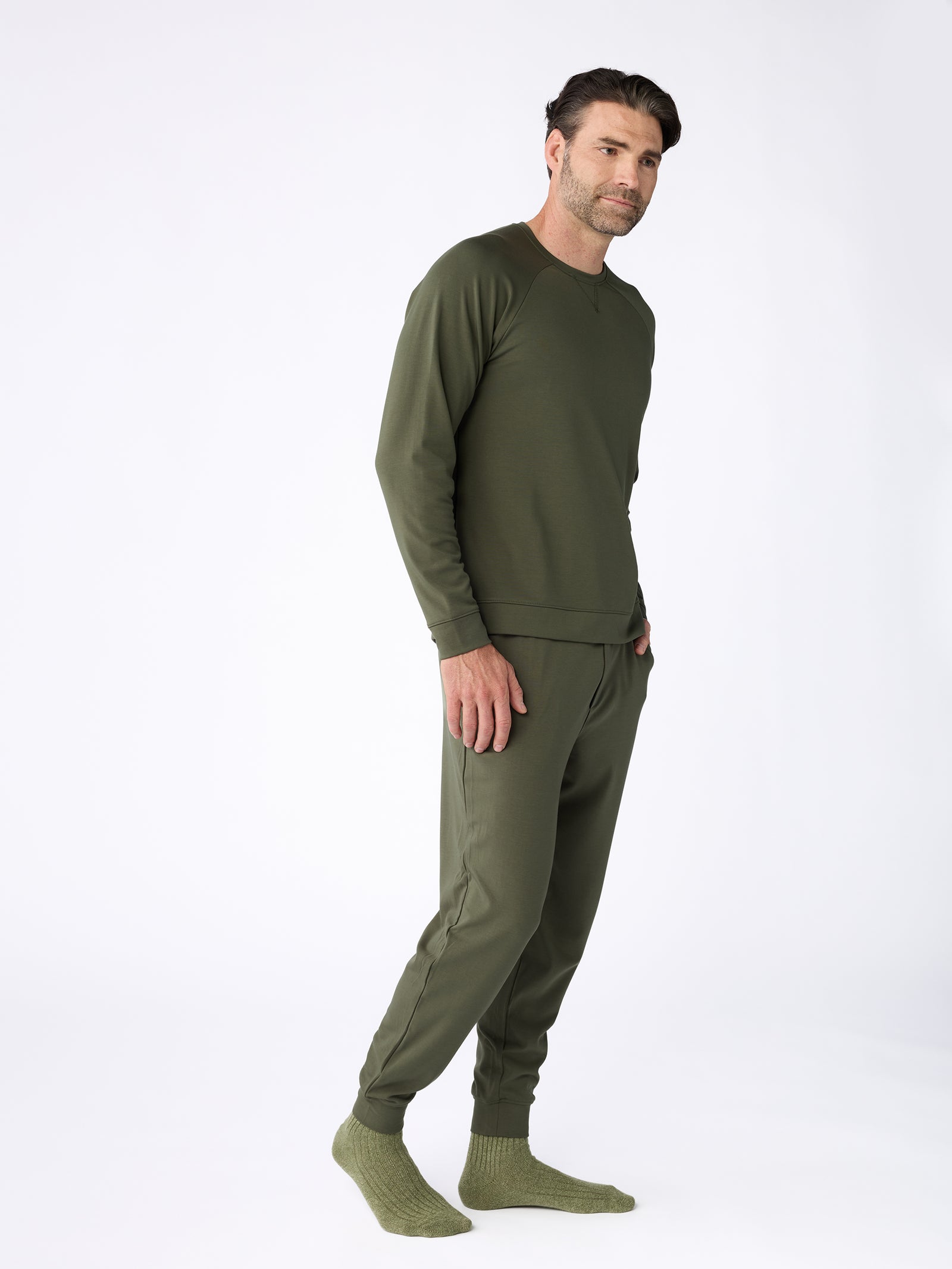 A person is wearing a matching olive green loungewear set from Cozy Earth, featuring the Men's Ultra-Soft Bamboo Pullover Crew and pants, paired with green socks. They stand against a plain white background in a relaxed pose, sporting a slight smile. 