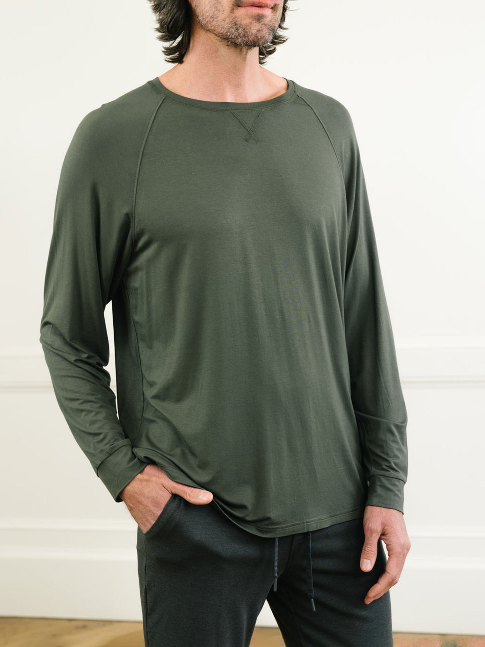 A person with shoulder-length hair stands with one hand in their pocket, wearing a dark green Cozy Earth Men's Stretch-Knit Long Sleeve Bamboo Pajama Set, which features raglan sleeves and appears lightweight. The background is a light-colored indoor setting with a wooden floor. 