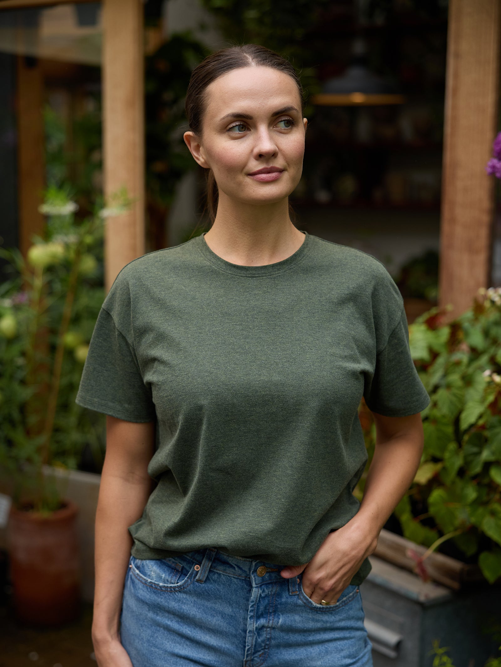 A woman with dark hair tied back is wearing the Women's All Day Tee in green from Cozy Earth, paired with blue jeans. She stands outdoors in a garden, framed by a wooden structure and various plants. With her left hand tucked into her pocket, she gazes slightly to the side. 