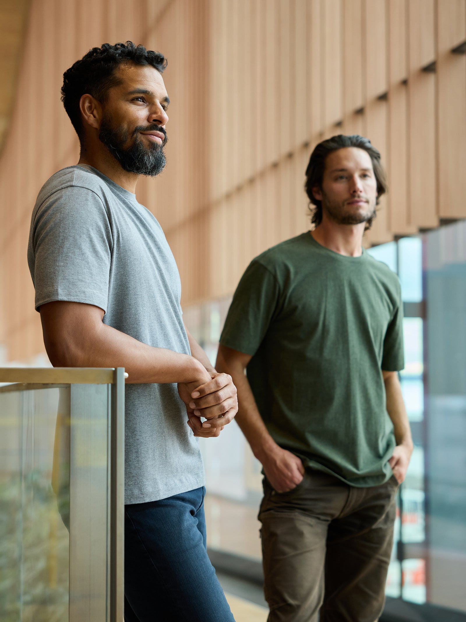 Two men stand side by side in a modern, well-lit indoor space with large windows. The man on the left wears a grey Men's All Day Tee from Cozy Earth; the man on the right wears a green Men's All Day Tee from Cozy Earth. Both are looking slightly off-camera in different directions, appearing relaxed. 