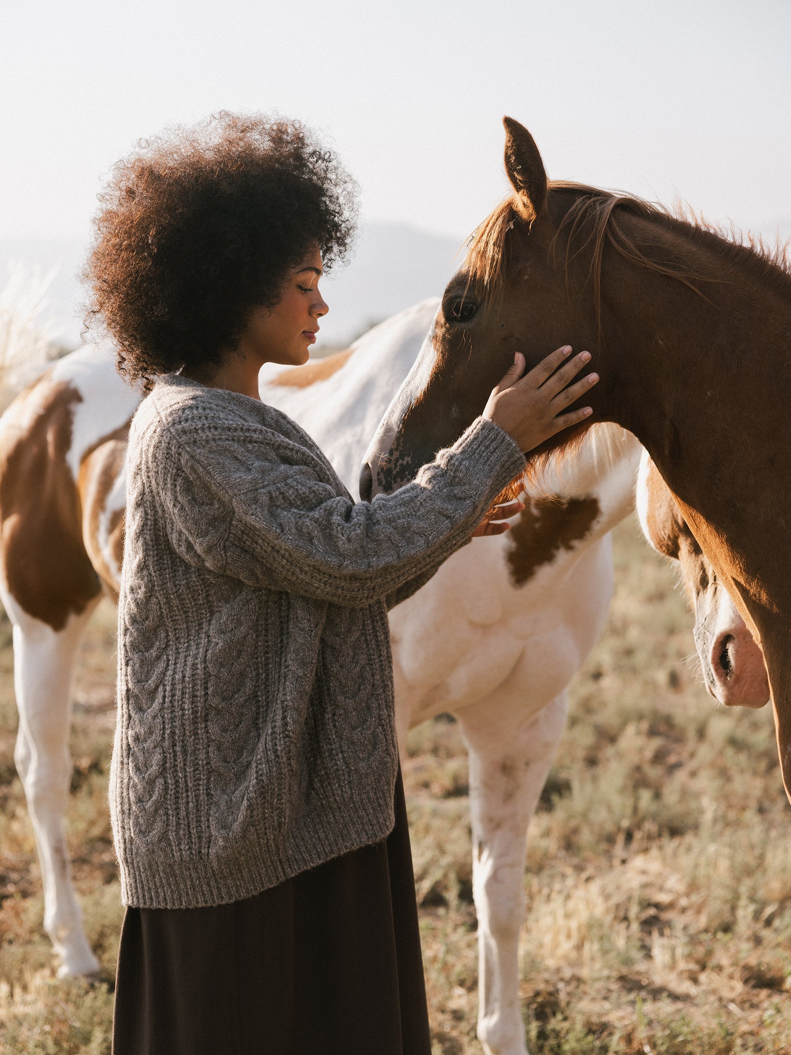 A person stands in a field, softly touching the nose of a horse with a white mane. They are wearing an Oversized Cable Knit Cardigan by Cozy Earth, and there are other horses in the background. The scene is peaceful and serene. 