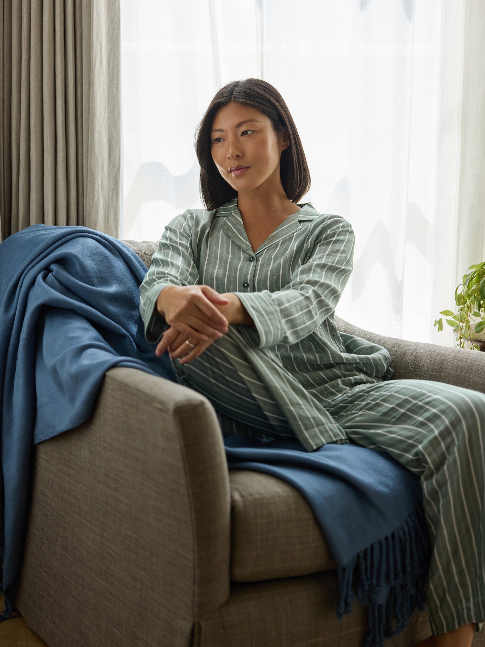 A person with long dark hair is sitting on a couch wearing green and white striped pajamas. A Bamboo Tassel Throw by Cozy Earth, in blue, is draped over the couch. The background features a window with sheer curtains and a small plant on the right. The person appears thoughtful and relaxed. 