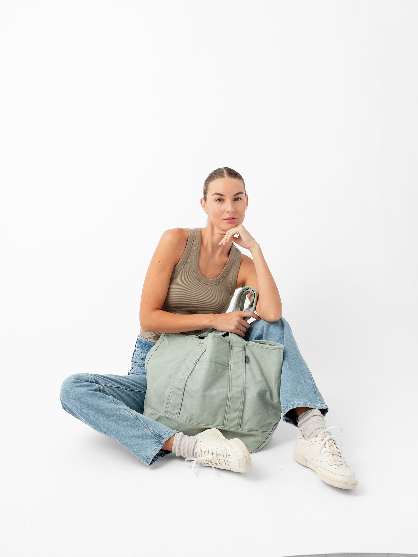 A woman with a pensive expression sits on the floor against a white background. She is wearing a beige tank top, blue jeans, and white sneakers. Resting her chin on her hand, she has a large Cozy Earth Waxed Canvas Tote in light green in front of her. 