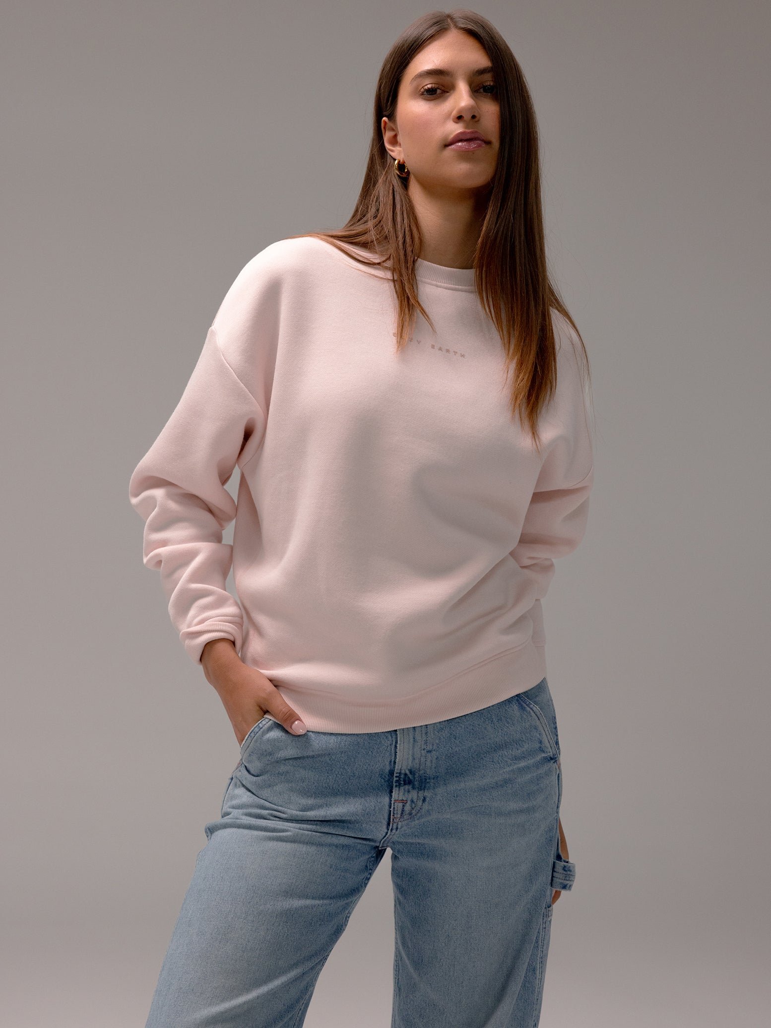 Someone with long hair models Cozy Earth's light pink Women's CityScape Crewneck paired with blue jeans, striking a casual pose with one hand in their pocket against a plain backdrop. 