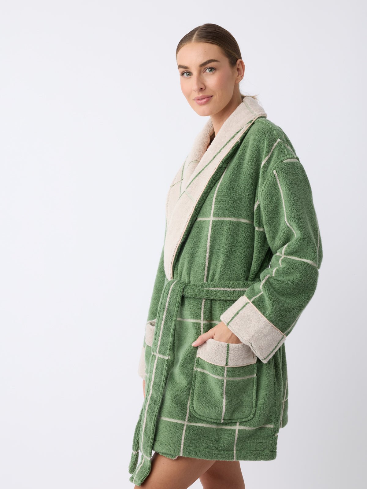 A person models the Cozy Earth Windowpane Resort Robe in green with a white grid, featuring large square pockets and a white collar. The robe is belted at the waist. They have a slight smile and their hair is pulled back against a plain background. 