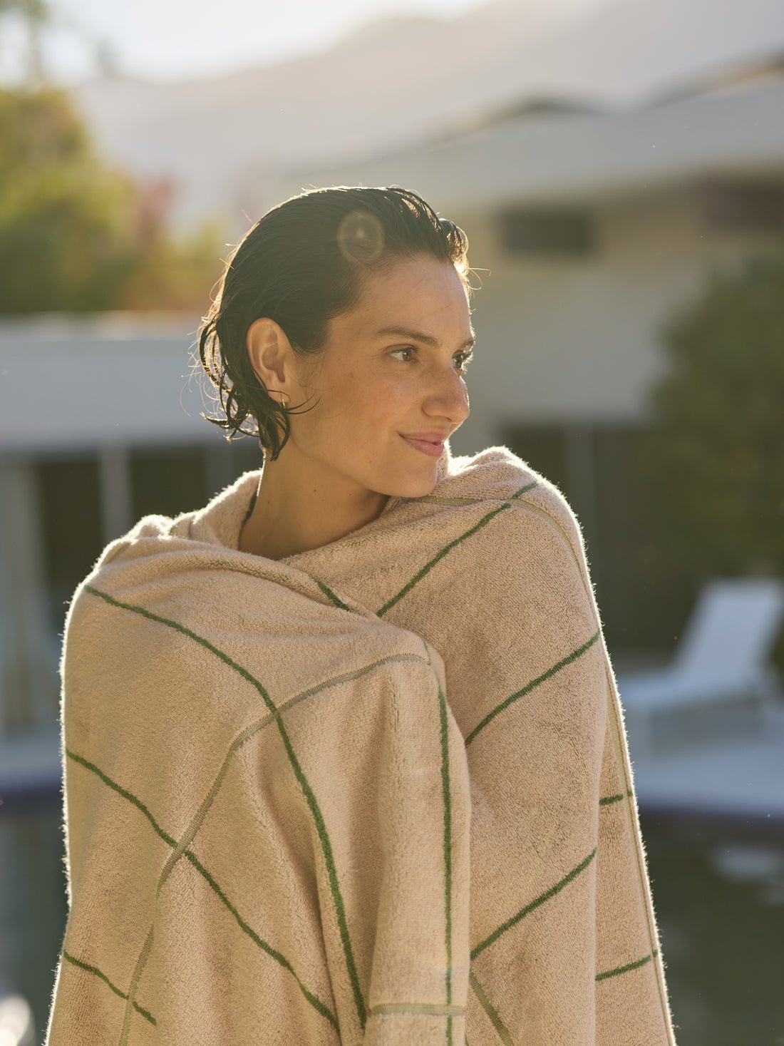 A person outdoors with short, wet hair wrapped in a Cozy Earth Windowpane Resort Towel. They stand by a pool, gazing sideways, against a backdrop of blurred trees and buildings. Soft sunlight creates a warm atmosphere. 