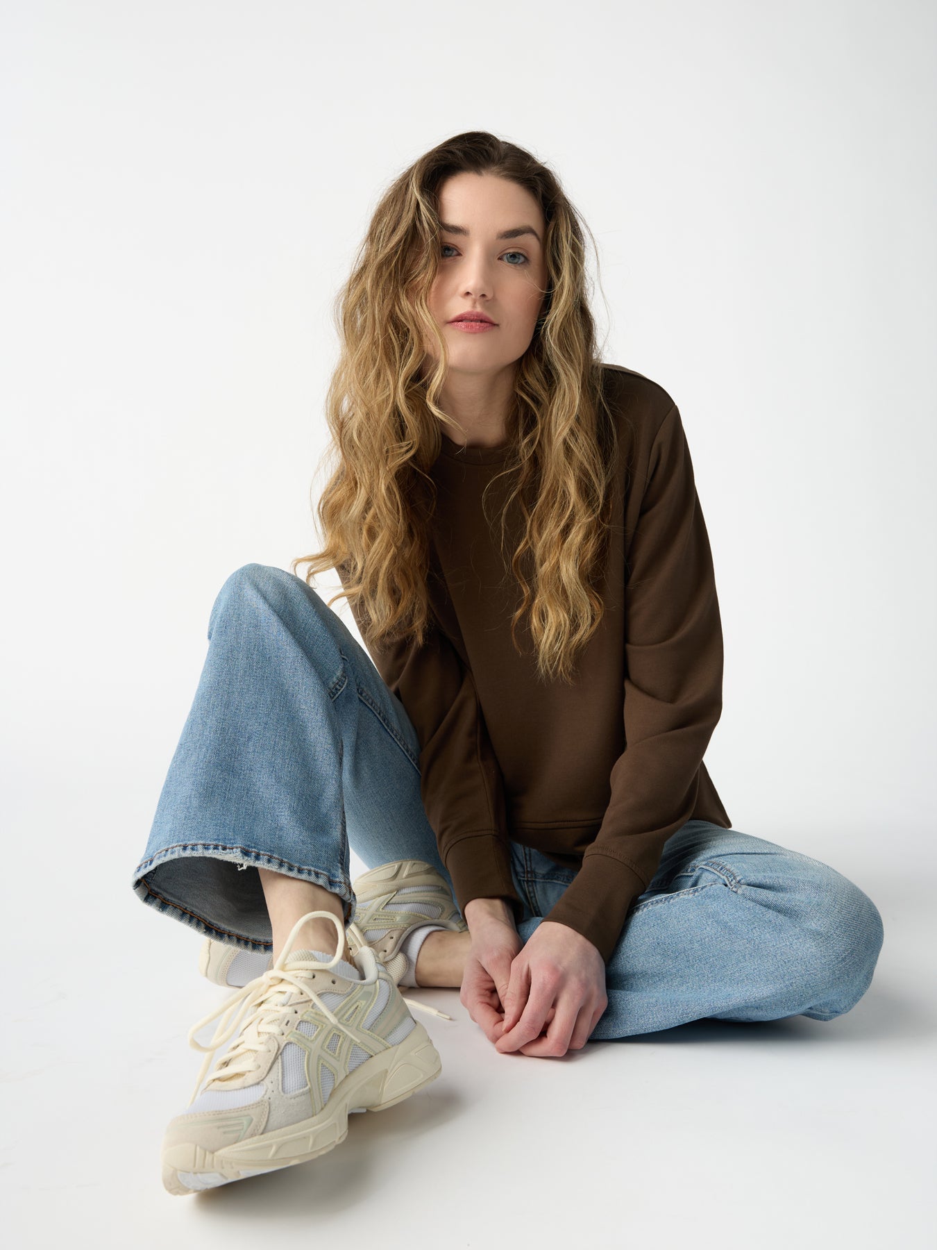 A person with long wavy hair sits relaxed on the ground. They are wearing a brown Women's StudioLite Crewneck by Cozy Earth, light blue jeans, and white sneakers. The plain white background gives a minimalist and clean look.