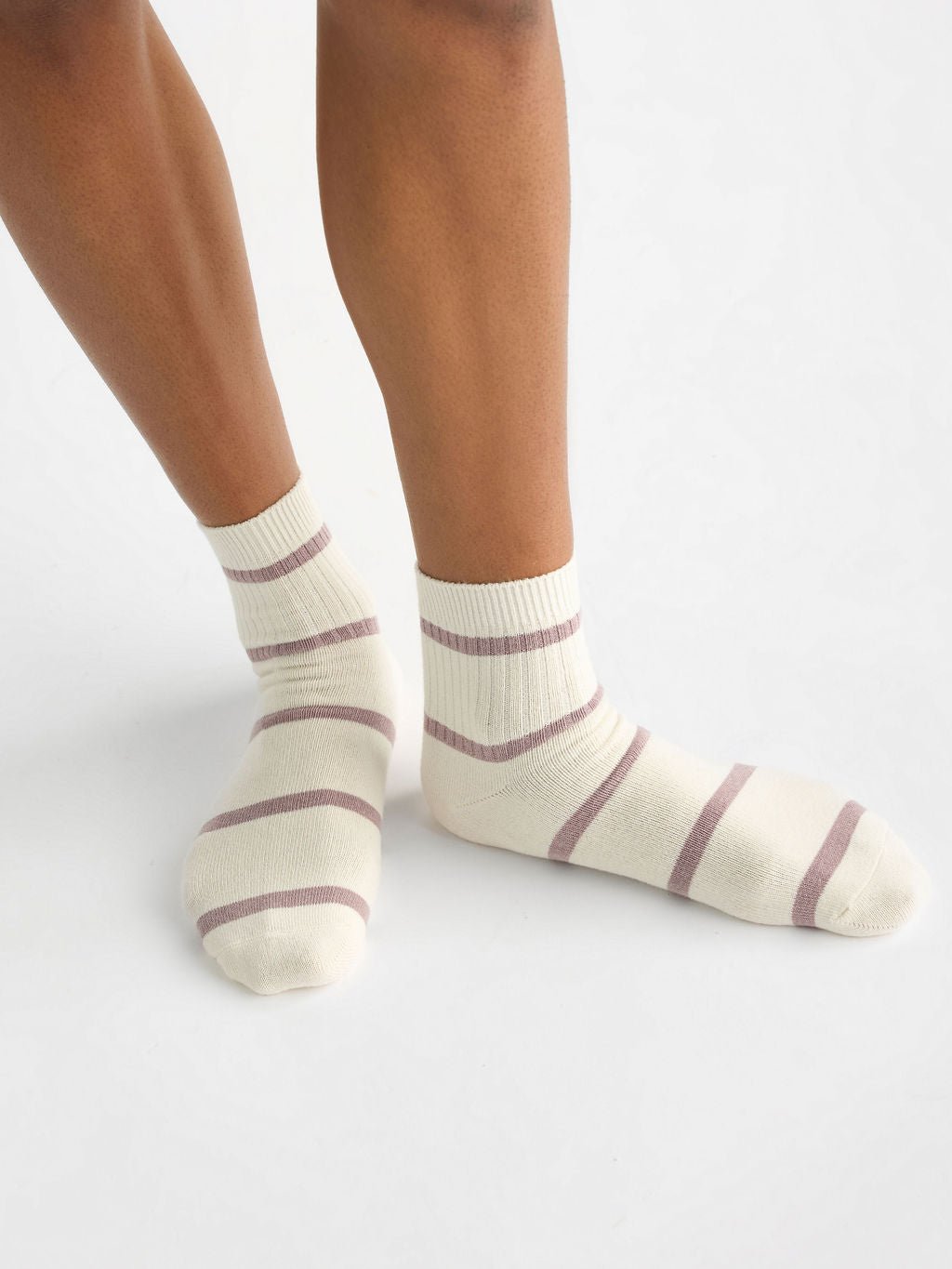 A person wearing Cozy Earth's Women's Sadie Sock, featuring cream color with purple stripes, is standing on a light gray surface. Only the lower legs and feet are visible. |Color:Parchment/Dusty Orchid