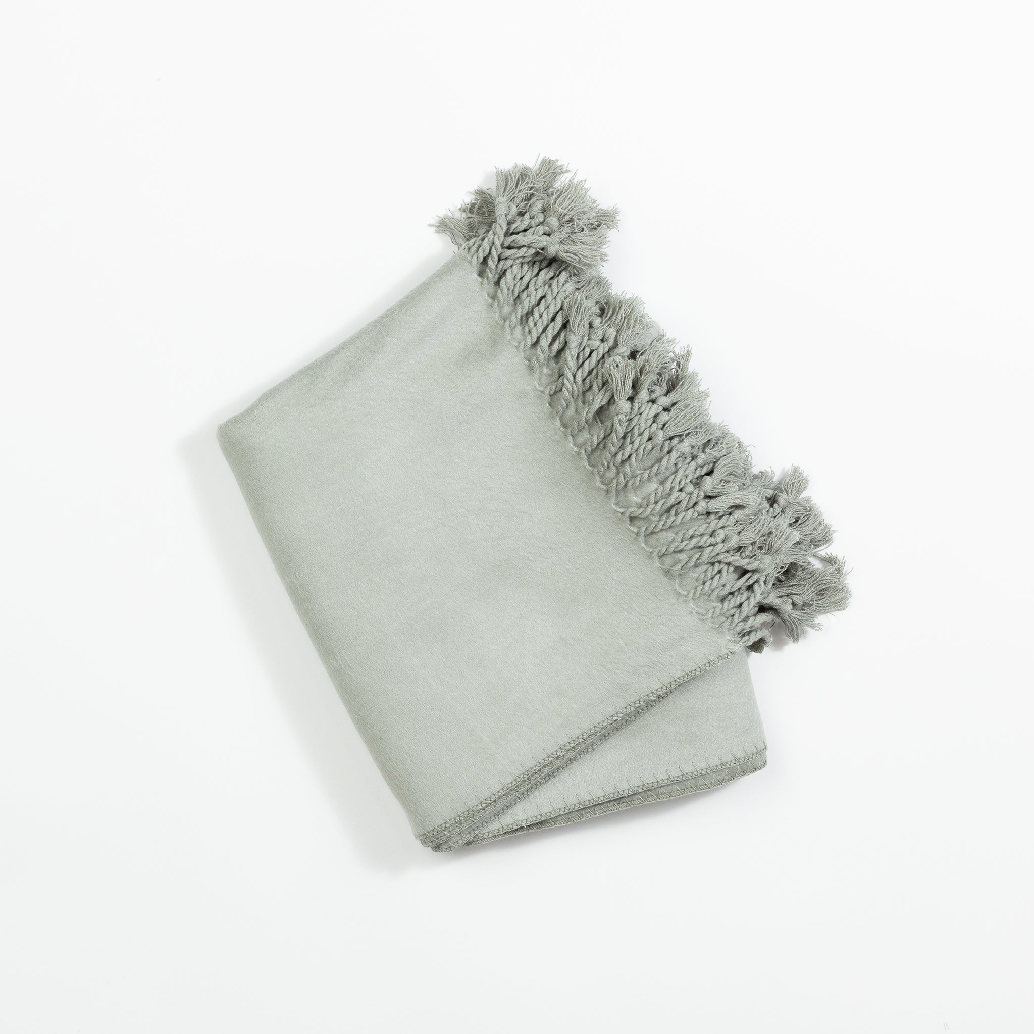 The Bamboo Tassel Throw by Cozy Earth, neatly folded in light gray with decorative tassels along one edge, is displayed on a plain white background. The tassels add a textured contrasting detail to the smooth fabric of the blanket. |Color:Sage