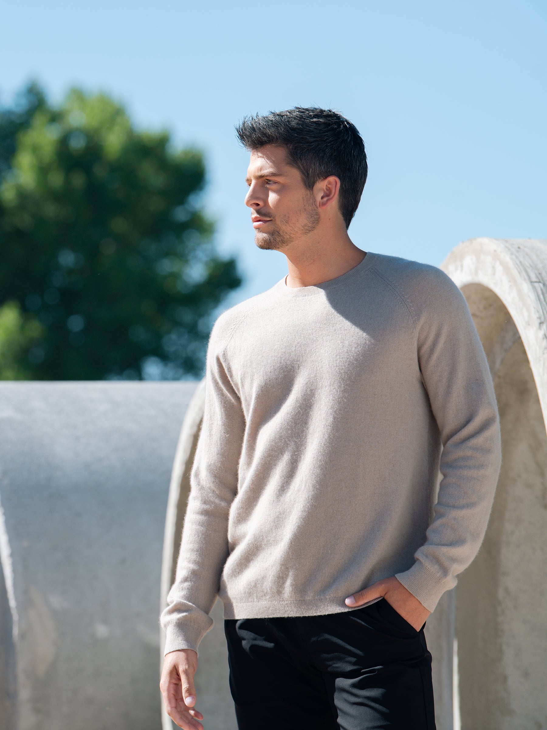 A man with short black hair is standing outdoors on a sunny day, wearing a light beige Men's Crewneck Sweater from Cozy Earth and black pants. His left hand is in his pants pocket, and he is looking to his left. Large circular concrete structures and a tree are in the background. |Color:Sandstone