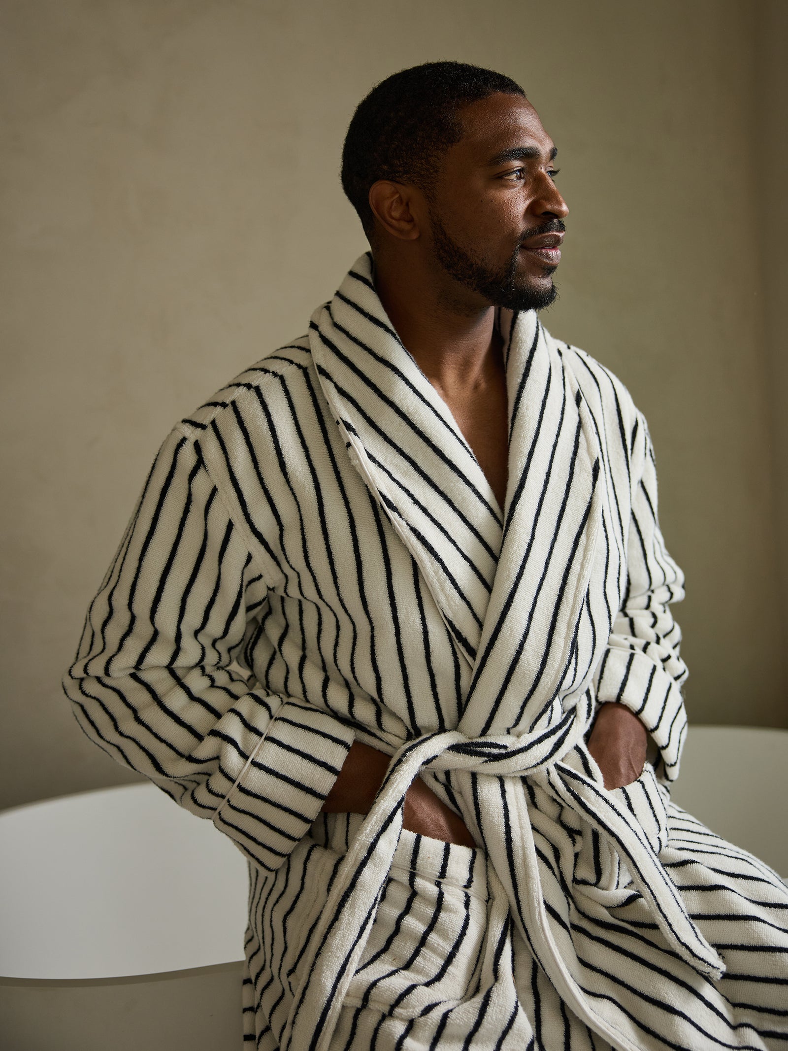 A man stands near a bathtub, dressed in the Cozy Earth Luxe Bath Robe, featuring a white design with black stripes. He gazes to the side with his hands tucked into the robe's pockets, set against a simple, neutral wall backdrop. 