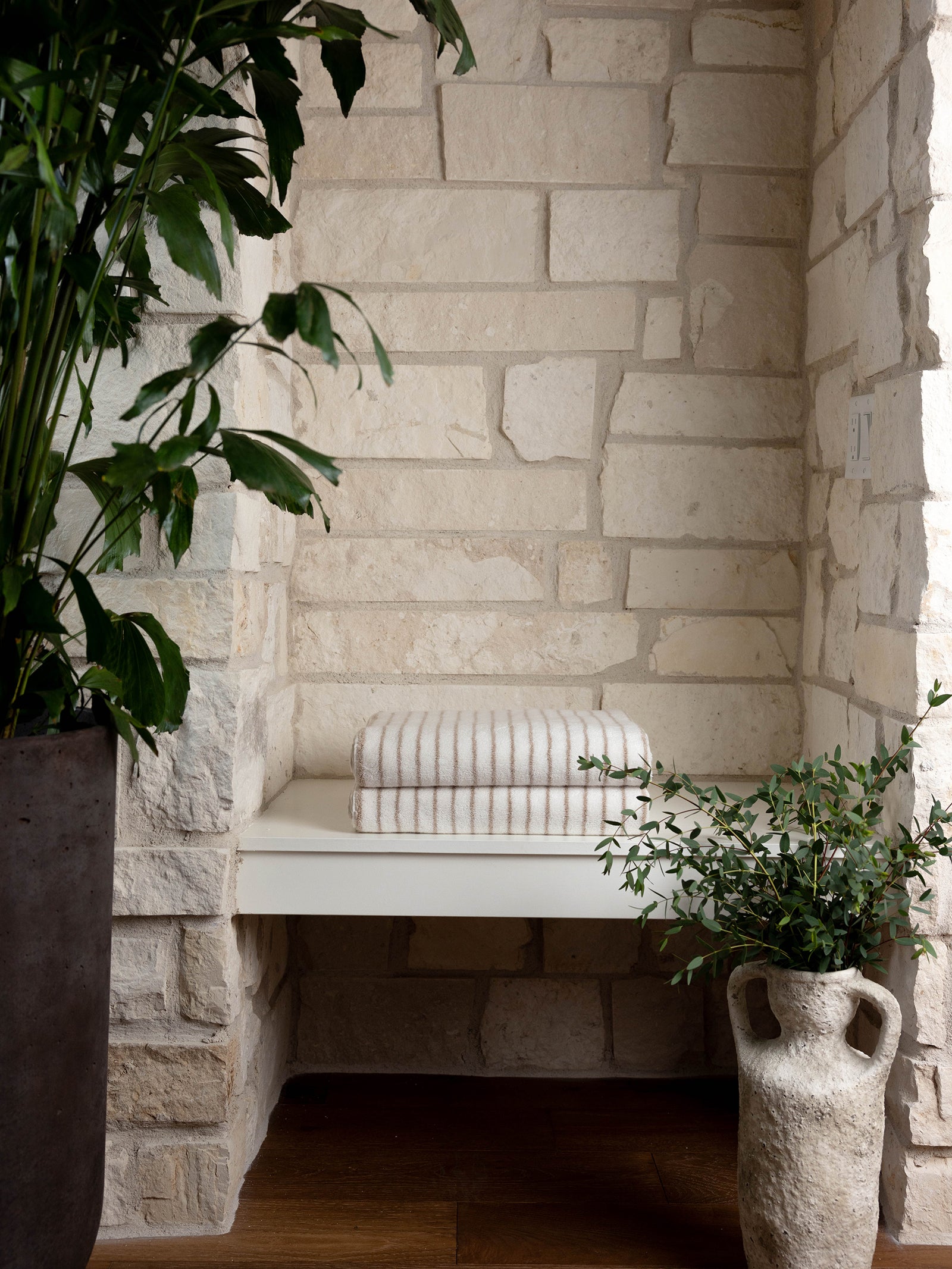 A cozy nook with a white stone wall features a minimalist cream-colored bench topped with two Luxe Bath Towels from Cozy Earth. The space is adorned with two lush green plants, one in a rustic gray pot and the other in an earthenware vase. Natural light highlights the serene setting. 