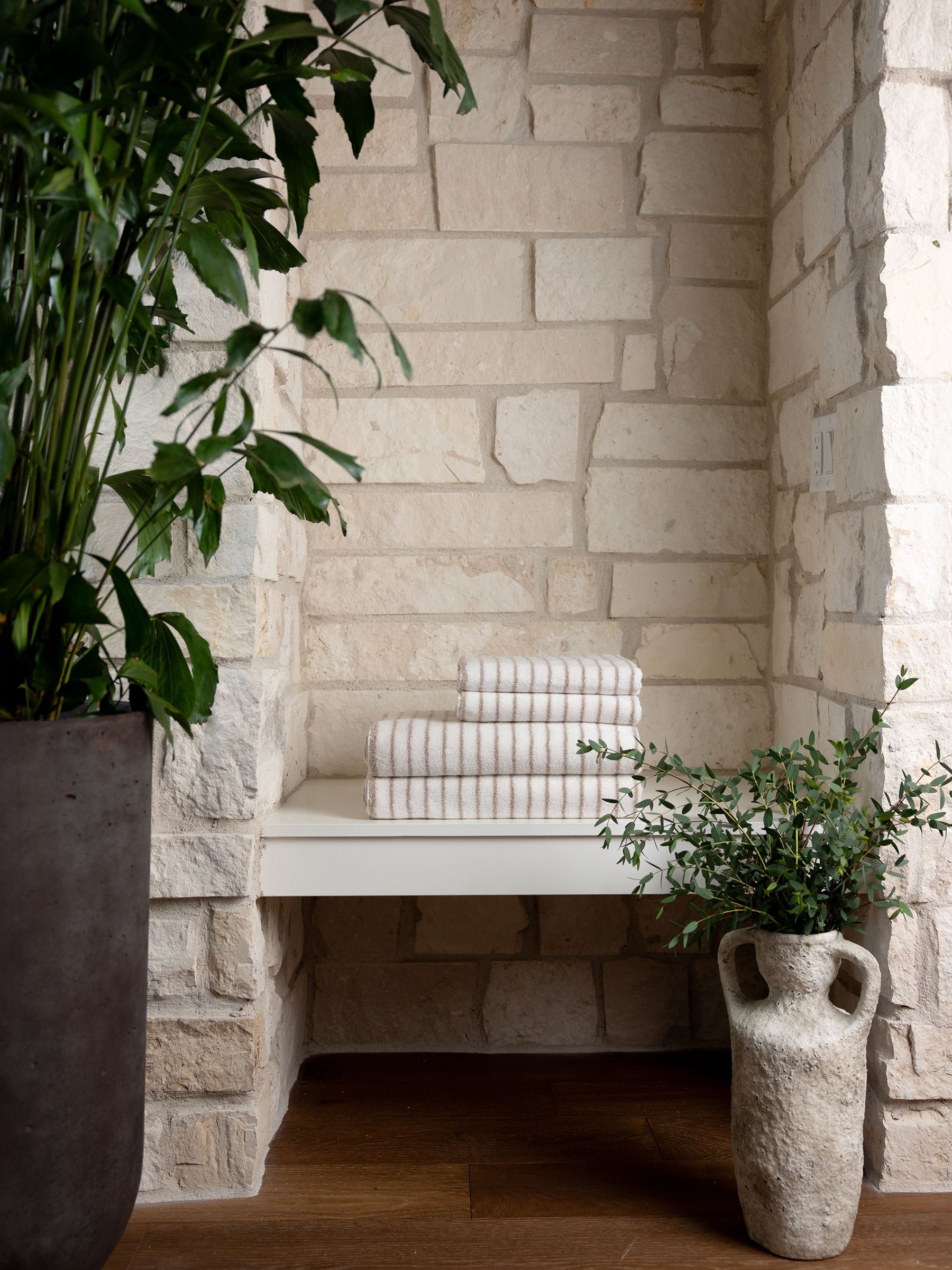 A serene corner with Luxe Hand Towels from Cozy Earth neatly stacked on a white floating shelf, surrounded by large potted plants. Walls feature light natural stone, and the floor is of dark wood, creating a harmonious blend of textures and natural colors. 