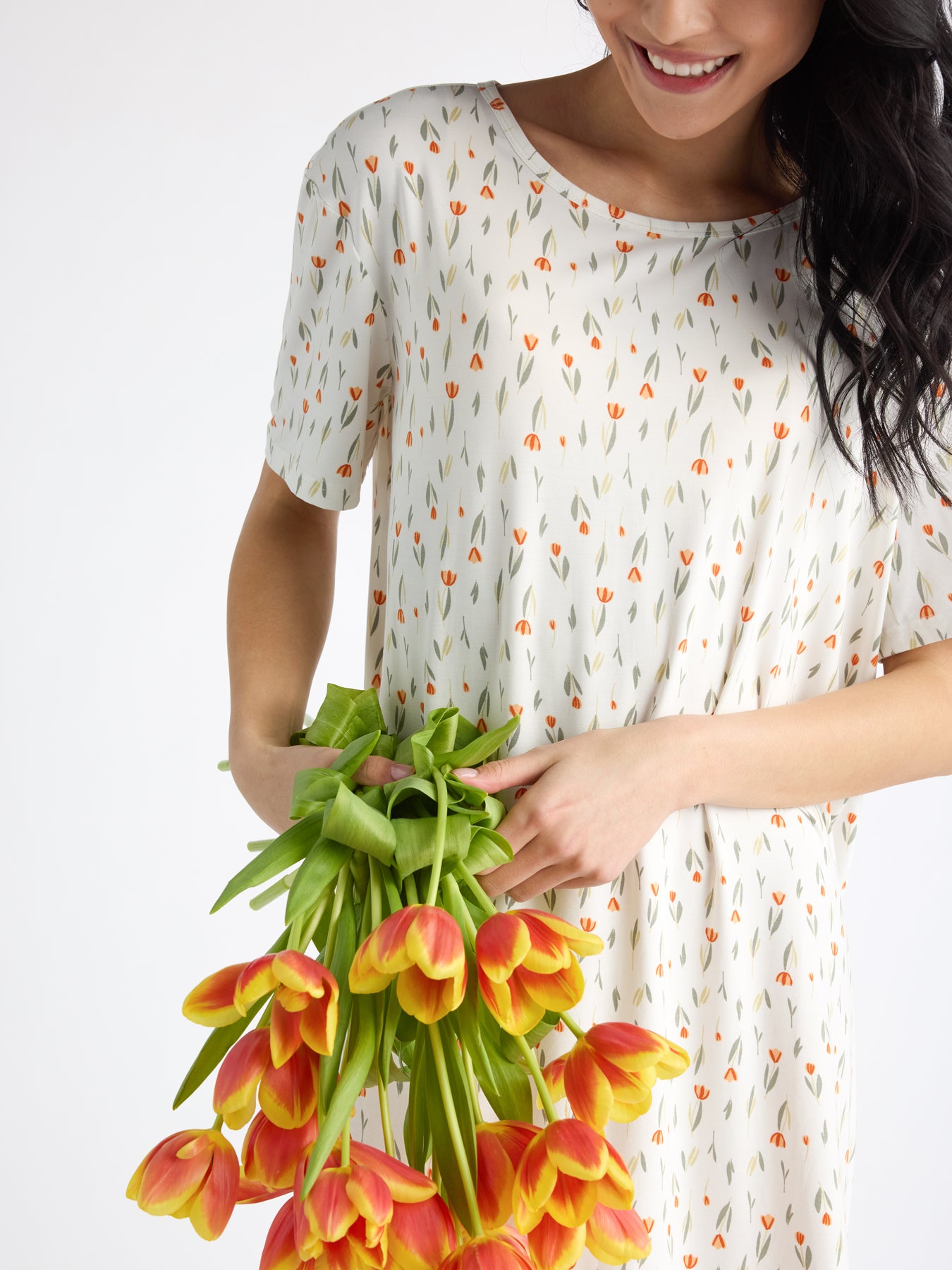 Wearing Cozy Earth's Women's Bamboo Stretch-Knit Sleep Dress with a delicate floral pattern, a person gently holds a bouquet of orange and yellow tulips. Their head is slightly lowered as they smile softly against the plain backdrop. 