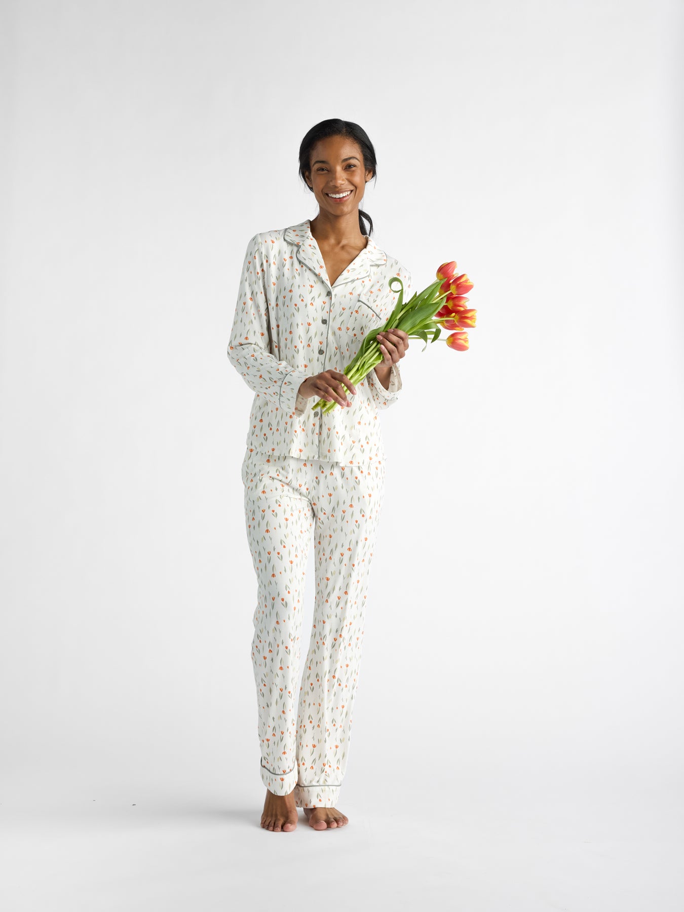 Standing barefoot against a plain background, the person wears Cozy Earth Women's Bamboo Stretch-Knit Long Sleeve Pajama Top with a colorful pattern. They're smiling and holding a bunch of orange tulips. 