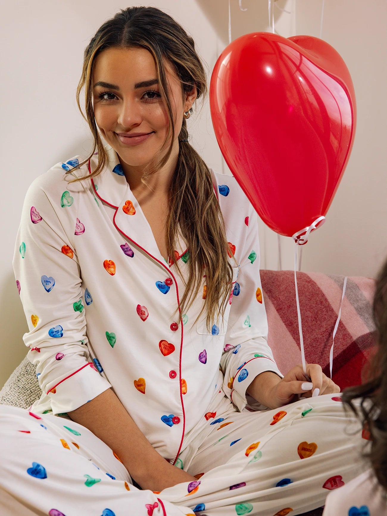 A woman in Cozy Earth's Women's Stretch-Knit Long Sleeve Bamboo Pajama Set sits cross-legged, holding a red heart-shaped balloon. They have long, brown hair and are smiling, surrounded by a cozy setting with a soft-looking blanket. 