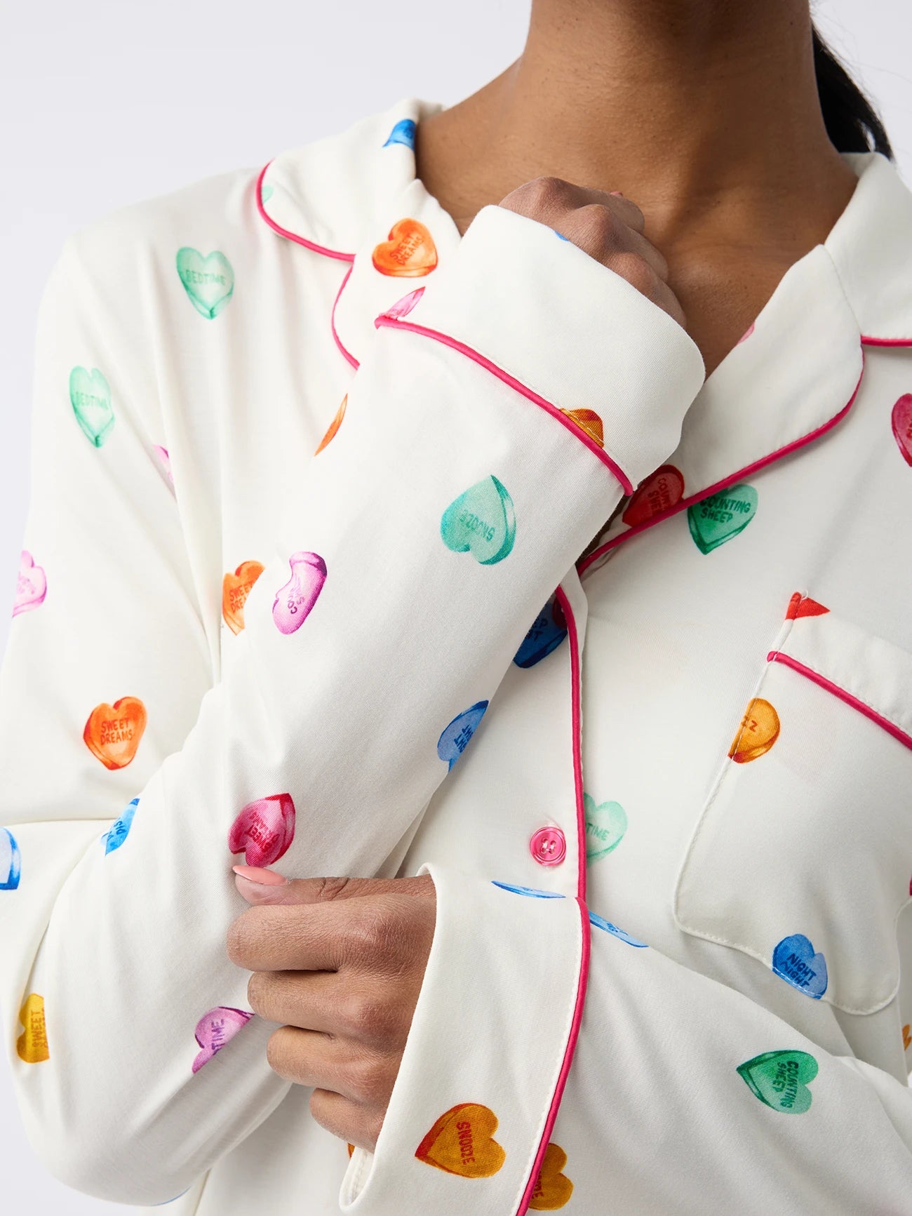 A person is wearing Cozy Earth Women's Long Sleeve Bamboo Pajama Top in Stretch-Knit, featuring a white color with colorful heart patterns, sweet messages on hearts, and pink piping. The person is partly visible with one hand touching the collar. 