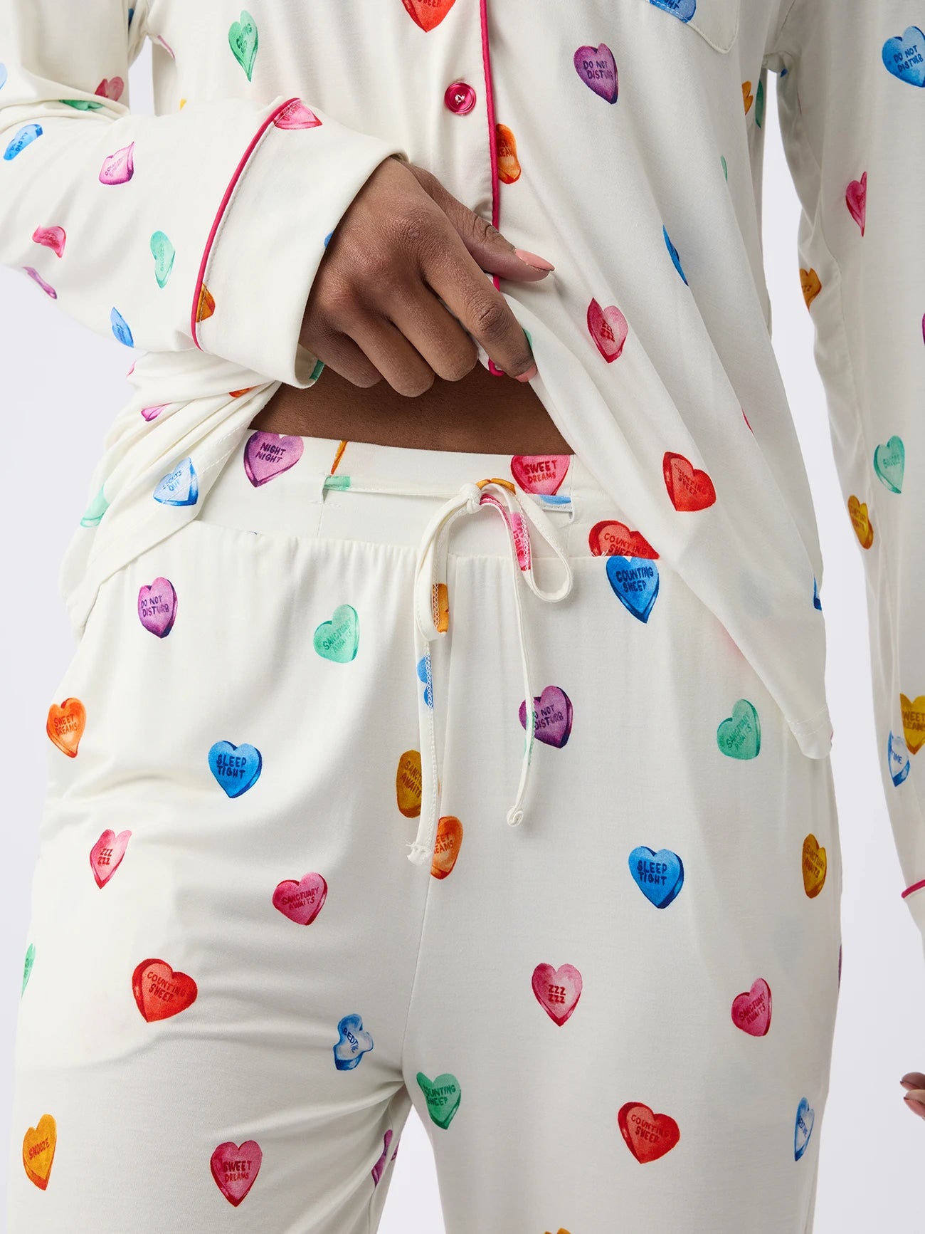 Someone is wearing Cozy Earth Women's Stretch-Knit Bamboo Pajama Pants in cream, featuring colorful heart designs with playful text. The top is lifted slightly to reveal matching drawstring pants against a plain white background. 