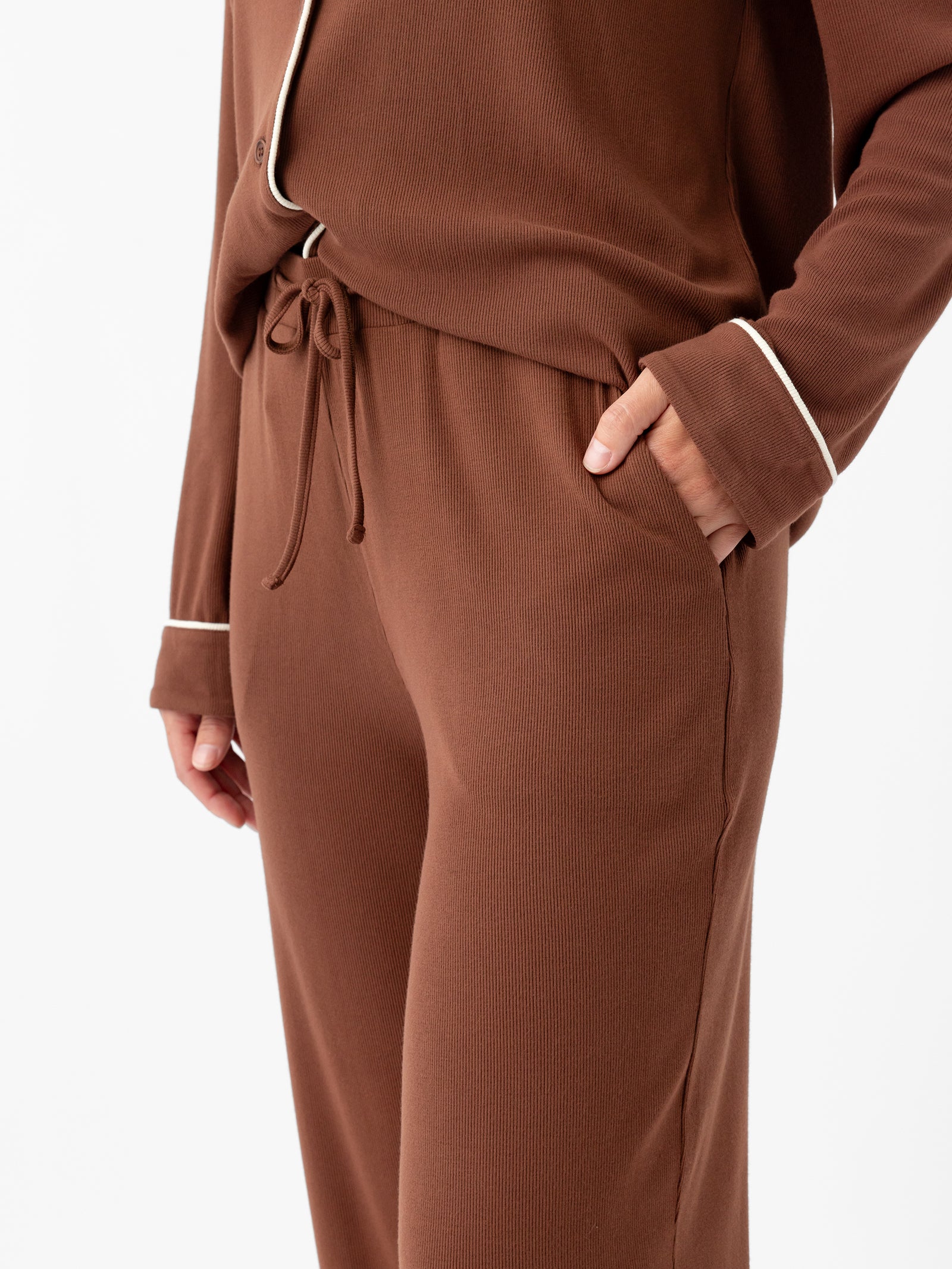 A person is shown from the neck down wearing a brown set from Cozy Earth, specifically the Women's Bamboo Rib Knit Classic Pajama Pant, which features white piping along the edges. The long-sleeved top has a loose, open front, while the pants include an elastic waistband with a drawstring. Their hands are casually placed in the side pockets. 