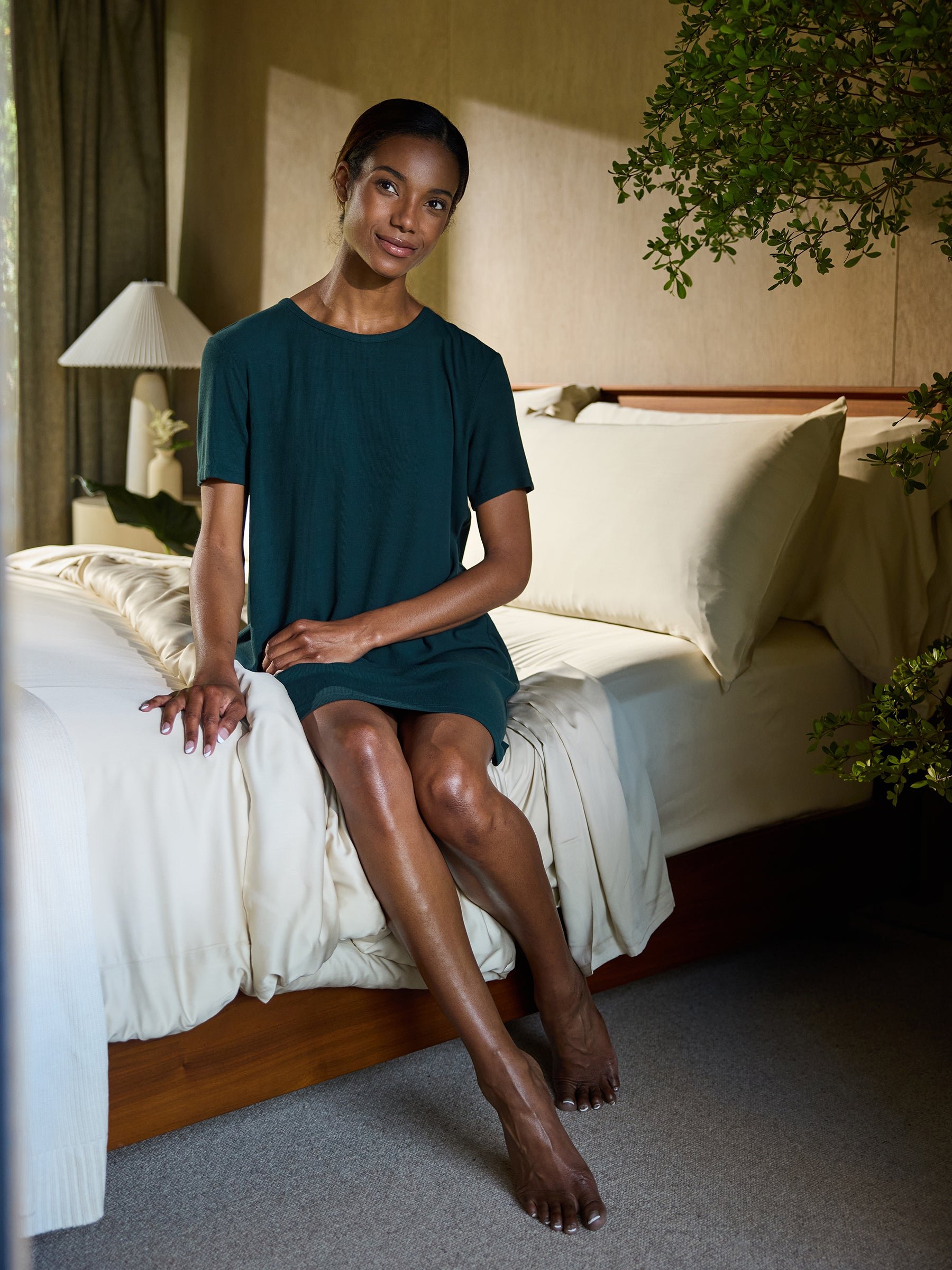 A woman wearing a Women's Bamboo Rib-Knit Boyfriend Sleep Dress by Cozy Earth sits on a bed with beige linens. She is resting her hands on the bed and looking at the camera with a slight smile. The bedroom is adorned with green indoor plants and has a cozy, well-lit ambiance. 