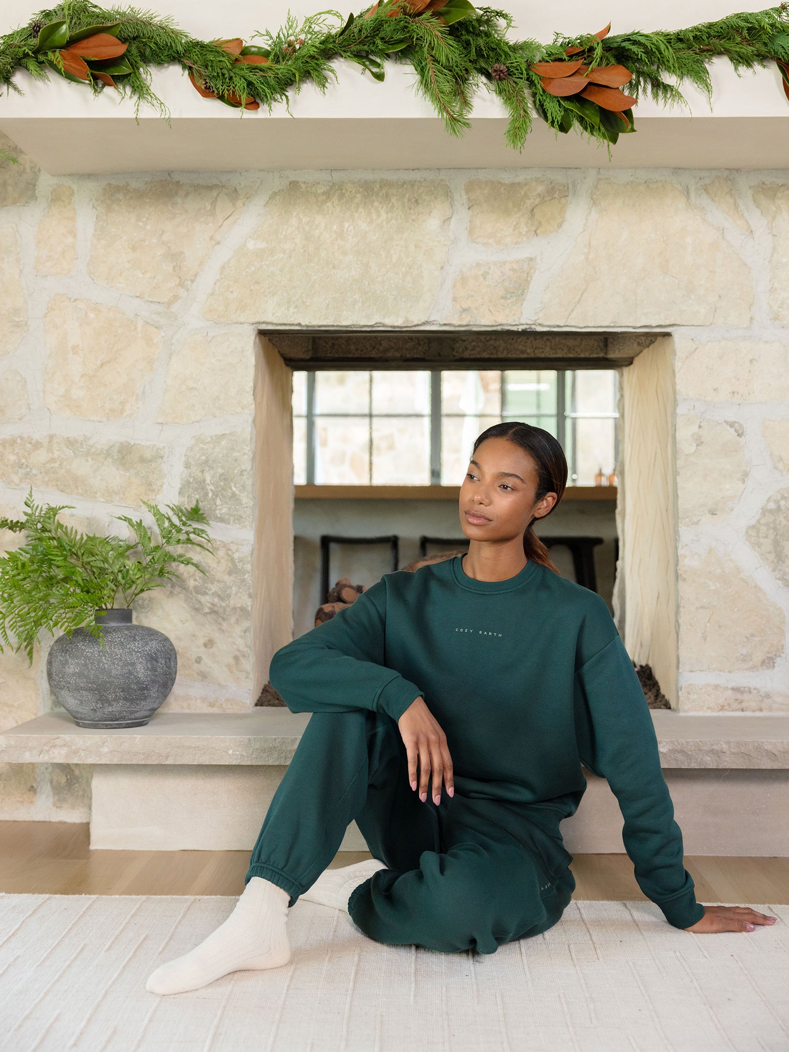 In a cozy stone-walled room, a woman sits on the floor wearing the Women's CityScape Crewneck by Cozy Earth in dark green, paired with matching sweatpants. A potted plant is beside her and festive greenery adorns the fireplace above. She appears relaxed and thoughtful. 