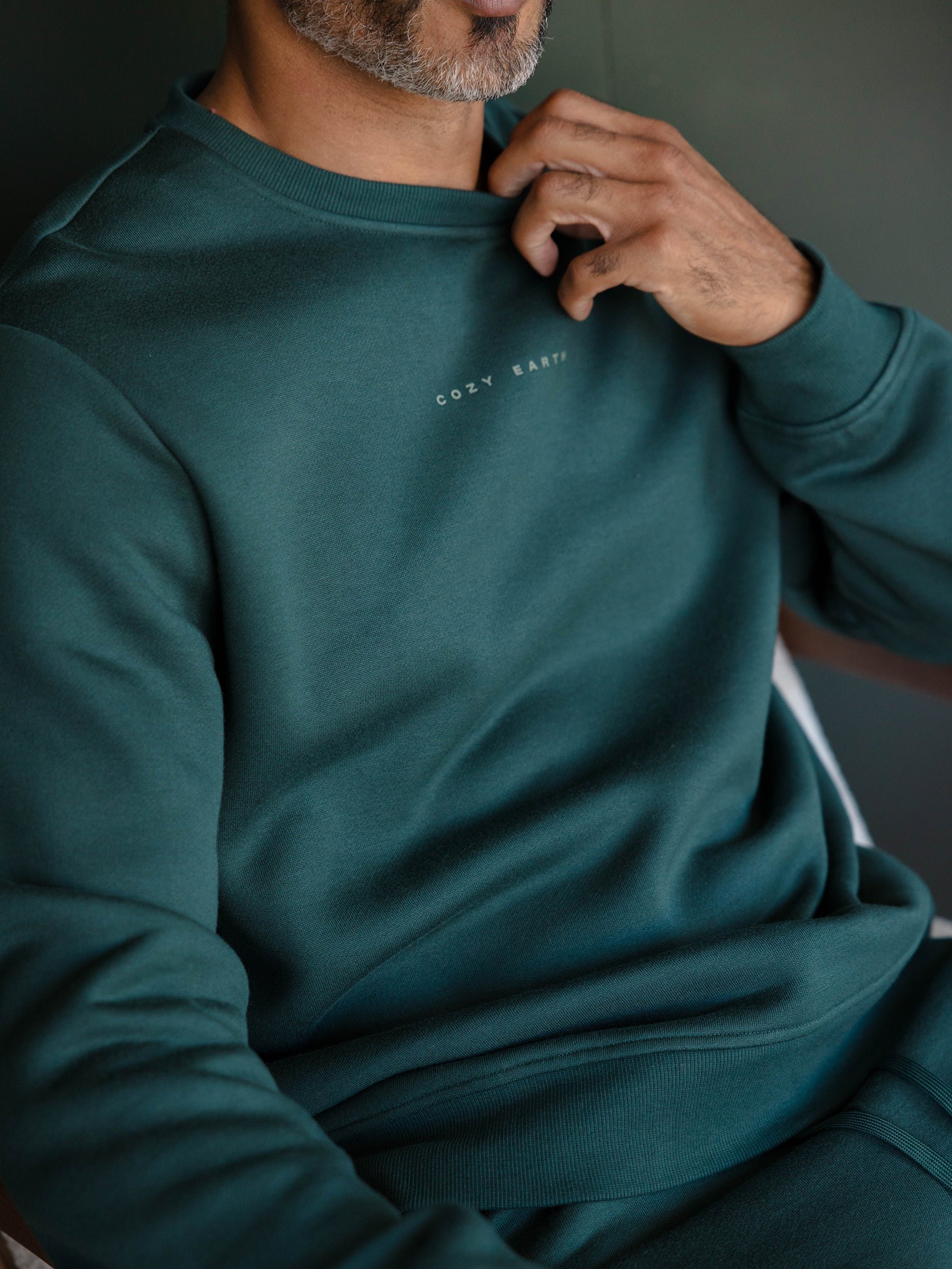A person wearing a dark green Men's CityScape Crewneck with "Cozy Earth" written on the chest, slightly tugging at the neckline. The image highlights the upper body and emphasizes the fabric texture. 