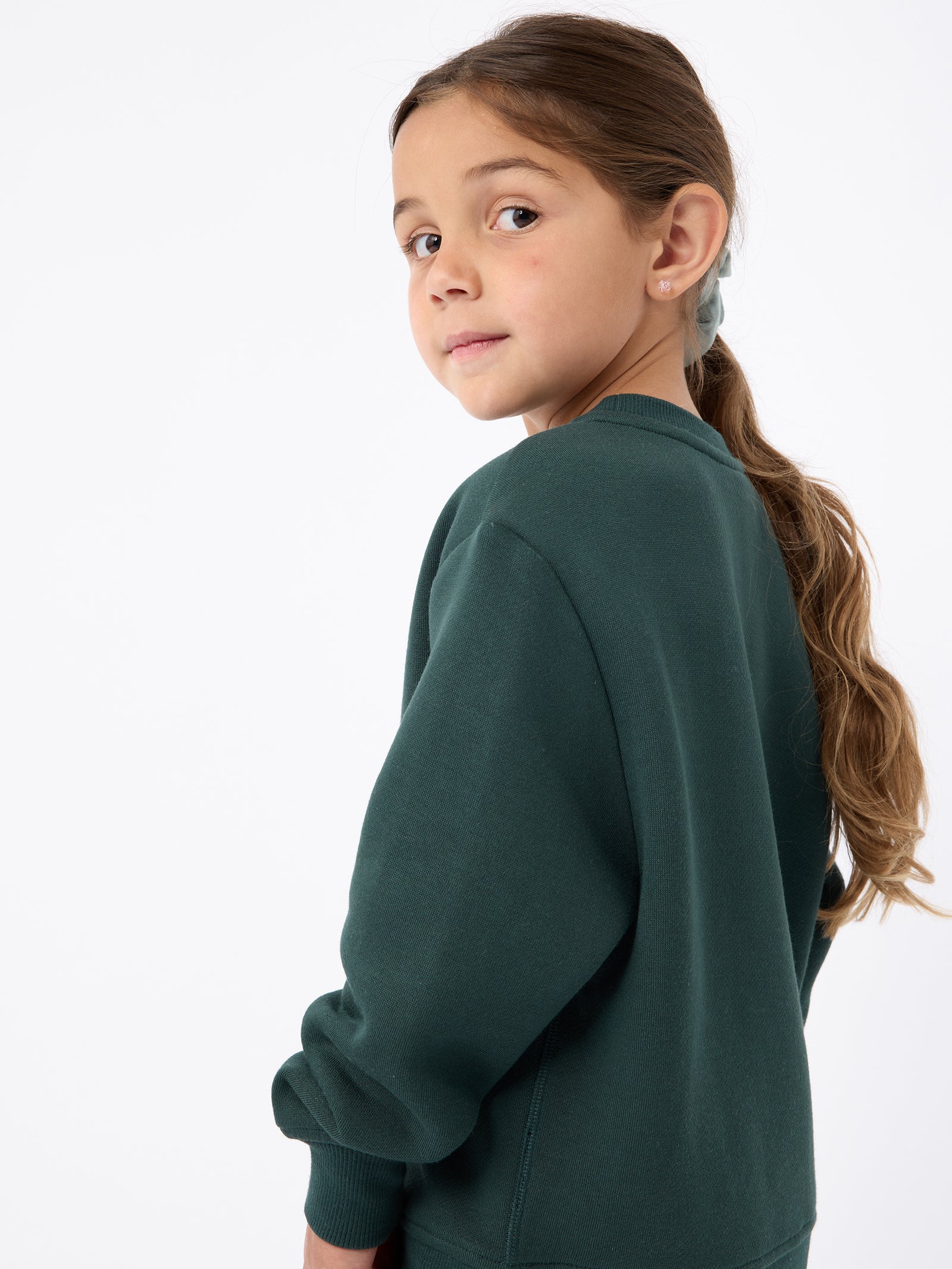 A young girl with long brown hair in a ponytail, wearing the Cozy Earth's Kid's CityScape Crewneck, looks over her shoulder against a plain white background. 