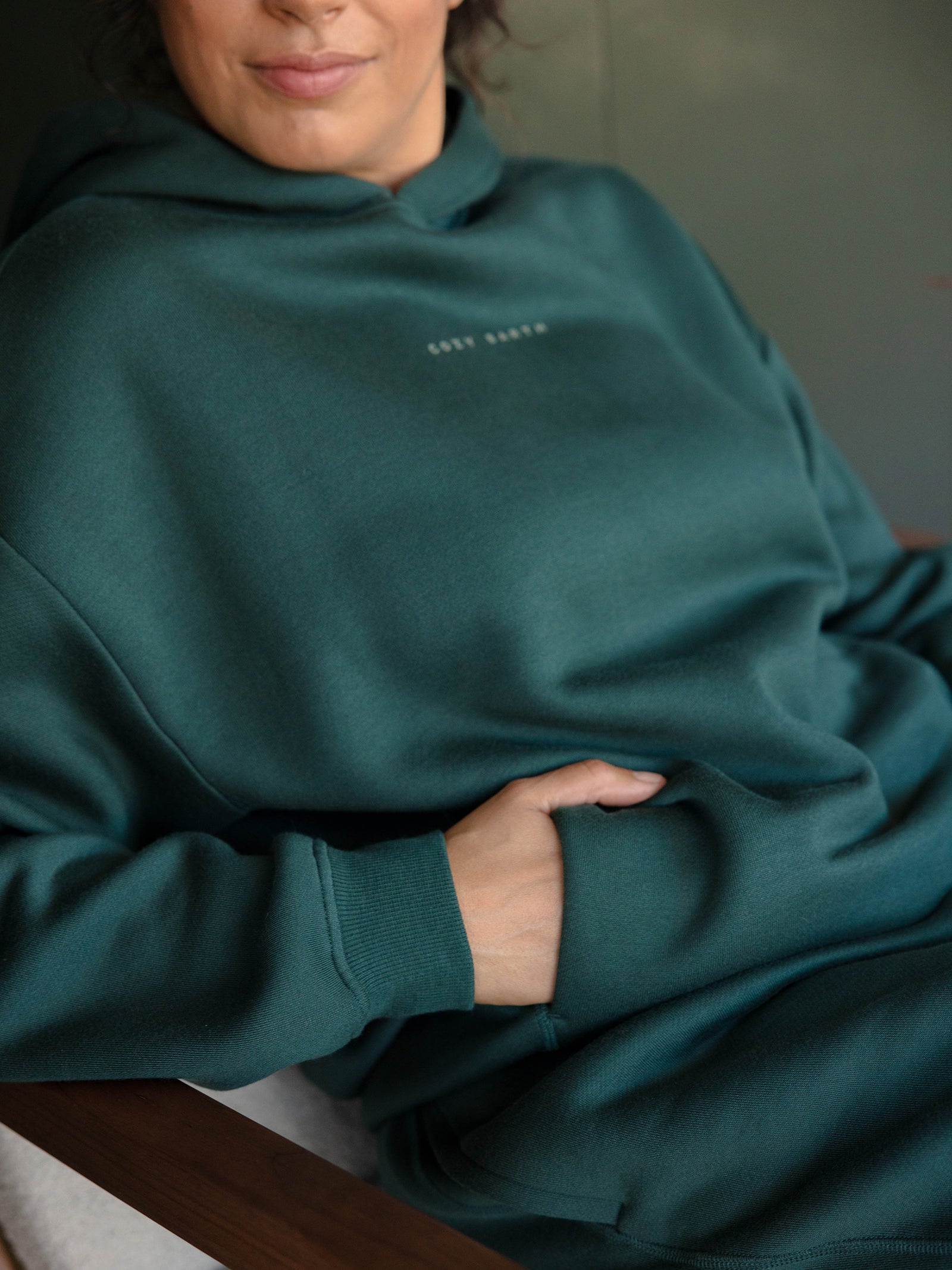 A person wearing a Cozy Earth Women's CityScape Hoodie in teal, featuring subtle chest text, sits in a chair with their left hand tucked into the hoodie's pocket against a softly blurred background. 