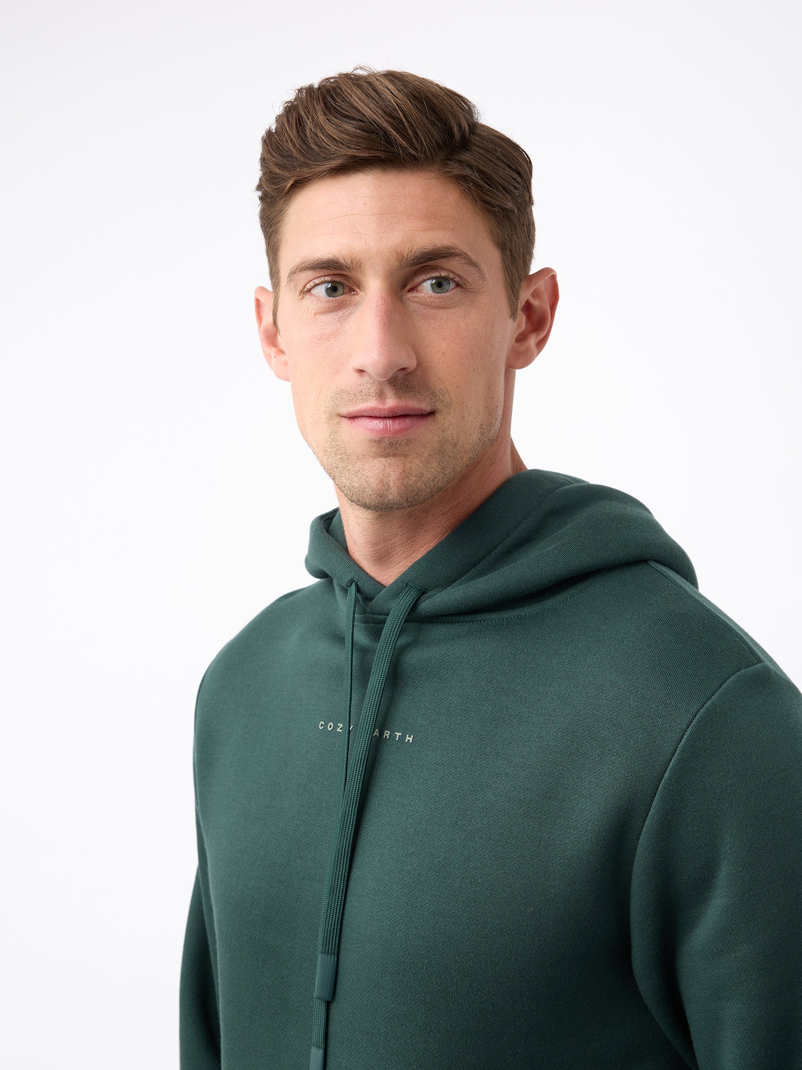 A person with short brown hair is wearing a Cozy Earth Men's CityScape Hoodie in dark green and looking to the side. The hoodie features subtle text on the chest, set against a plain white background. 