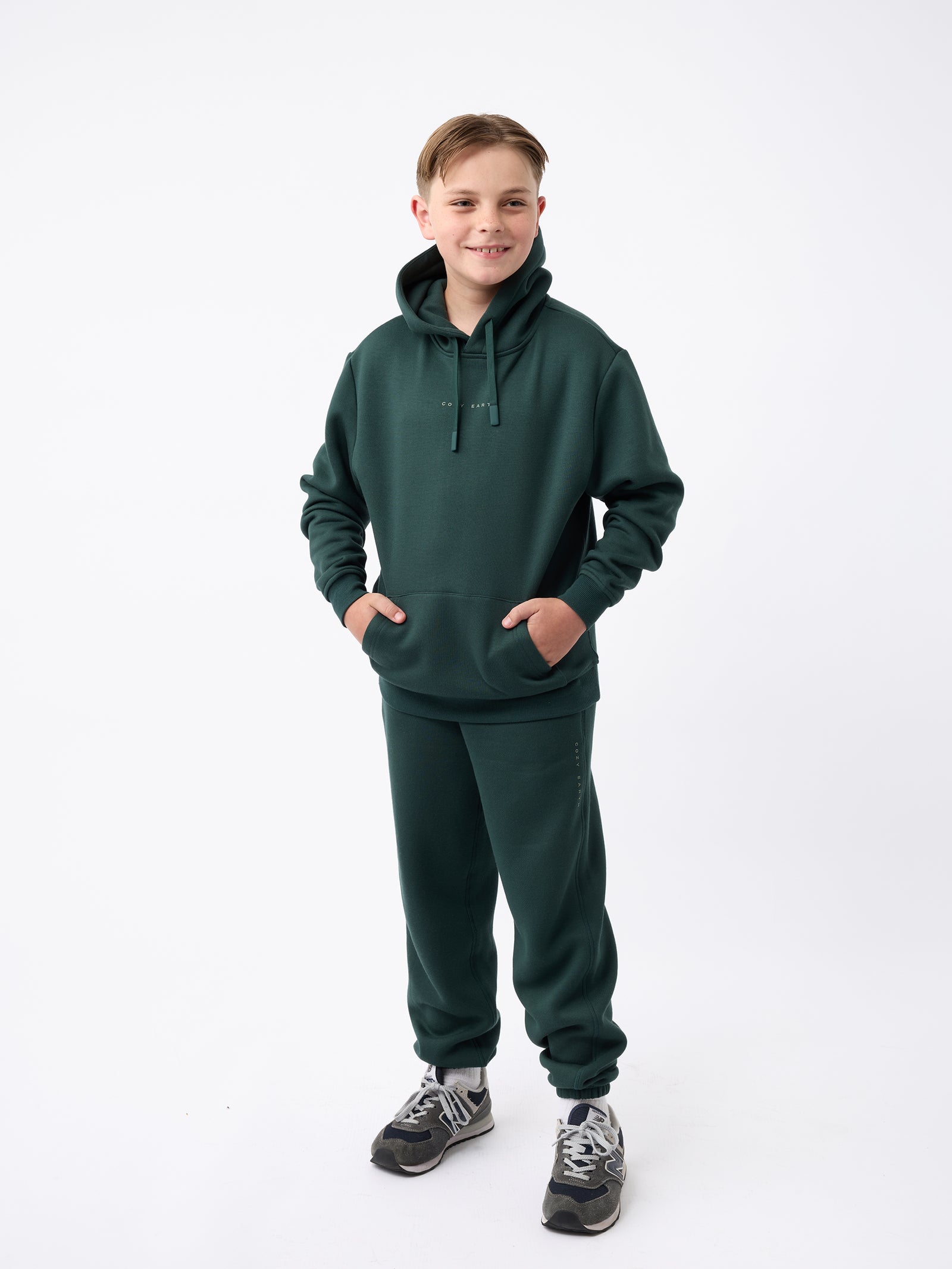 A young person stands smiling, dressed in a dark green Kid's CityScape Hoodie by Cozy Earth and matching sweatpants. Their hands are tucked in the hoodie pockets. They sport black and gray sneakers, all set against a plain white background. 