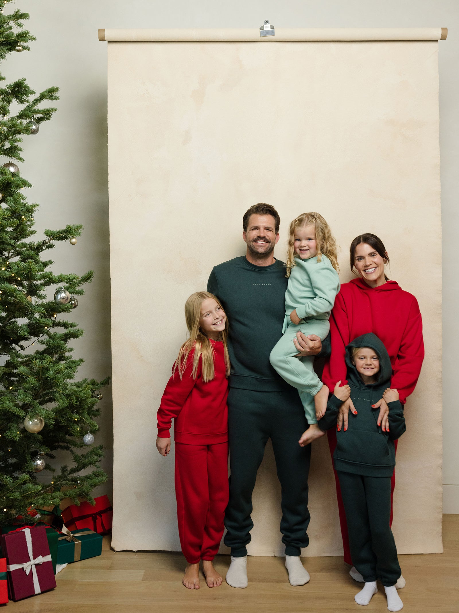 A family of five poses for a holiday photo with the father holding the youngest child. They are all dressed in festive red and green outfits from Cozy Earth. To the left, there's a decorated Christmas tree with presents, set against a beige backdrop. 