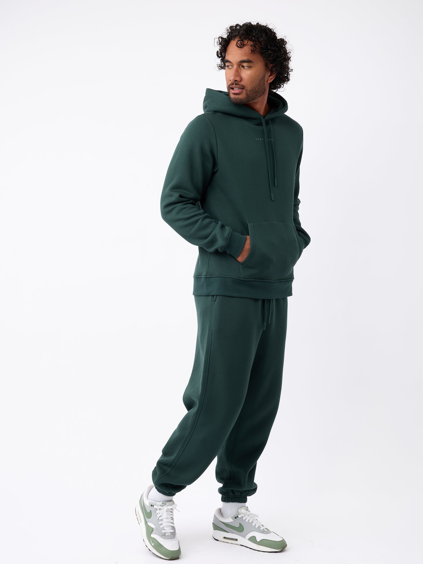 A man stands in a relaxed pose wearing a dark green hoodie paired with Cozy Earth's Men's CityScape Sweatpant. He has one hand in his pocket while looking to the side, and he complements his outfit with white and green sneakers against a white background. 