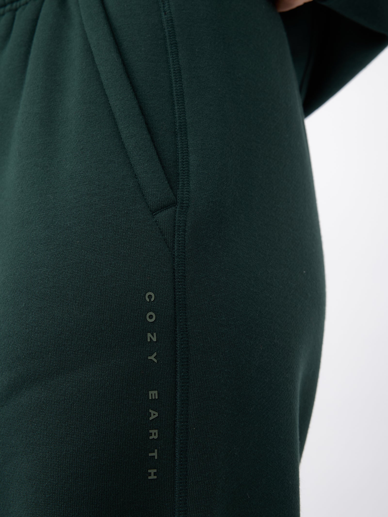 A close-up of a person wearing Women's CityScape Wide Leg Pant in dark green shows the brand name "Cozy Earth" printed vertically along the seam near the pocket. 