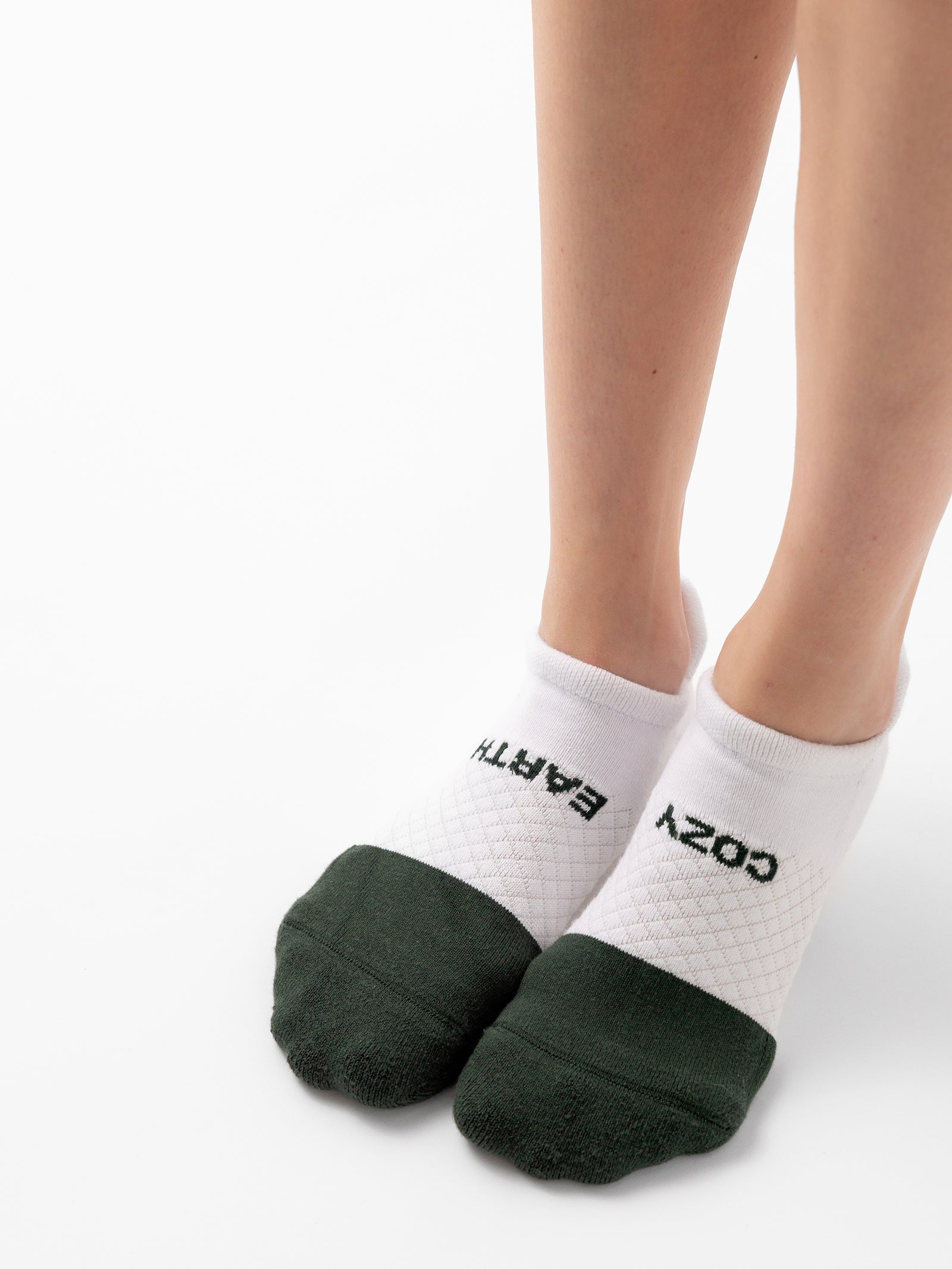 Close-up of a person's feet wearing white ankle socks from the "Essential Ankle Sock 2-Pack" by Cozy Earth. The socks feature dark green toes and heels, with "COZY EARTH" written on them—"COZY" on the right foot and "EARTH" on the left foot. The background is plain white. 