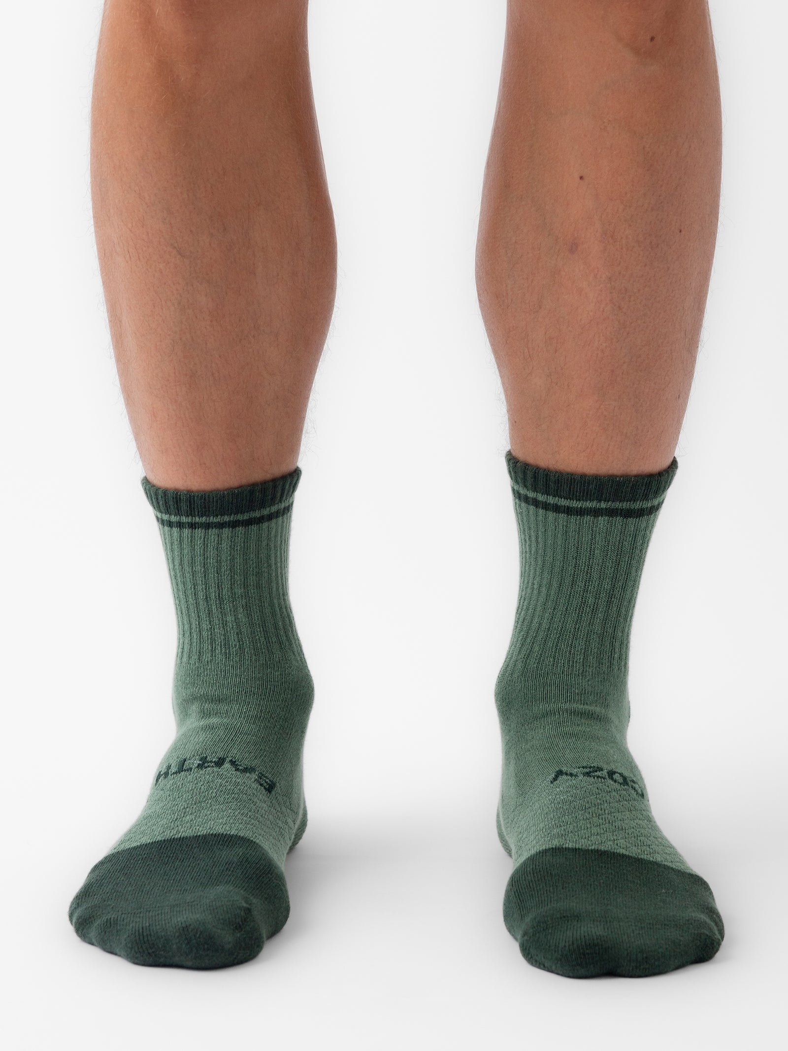 Close-up photo of a person's lower legs wearing the Essential Quarter Sock from Cozy Earth in light green with dark green toes, heels, and cuffs. The ribbed texture of the socks is clearly visible as the person stands barefoot on a white background. 