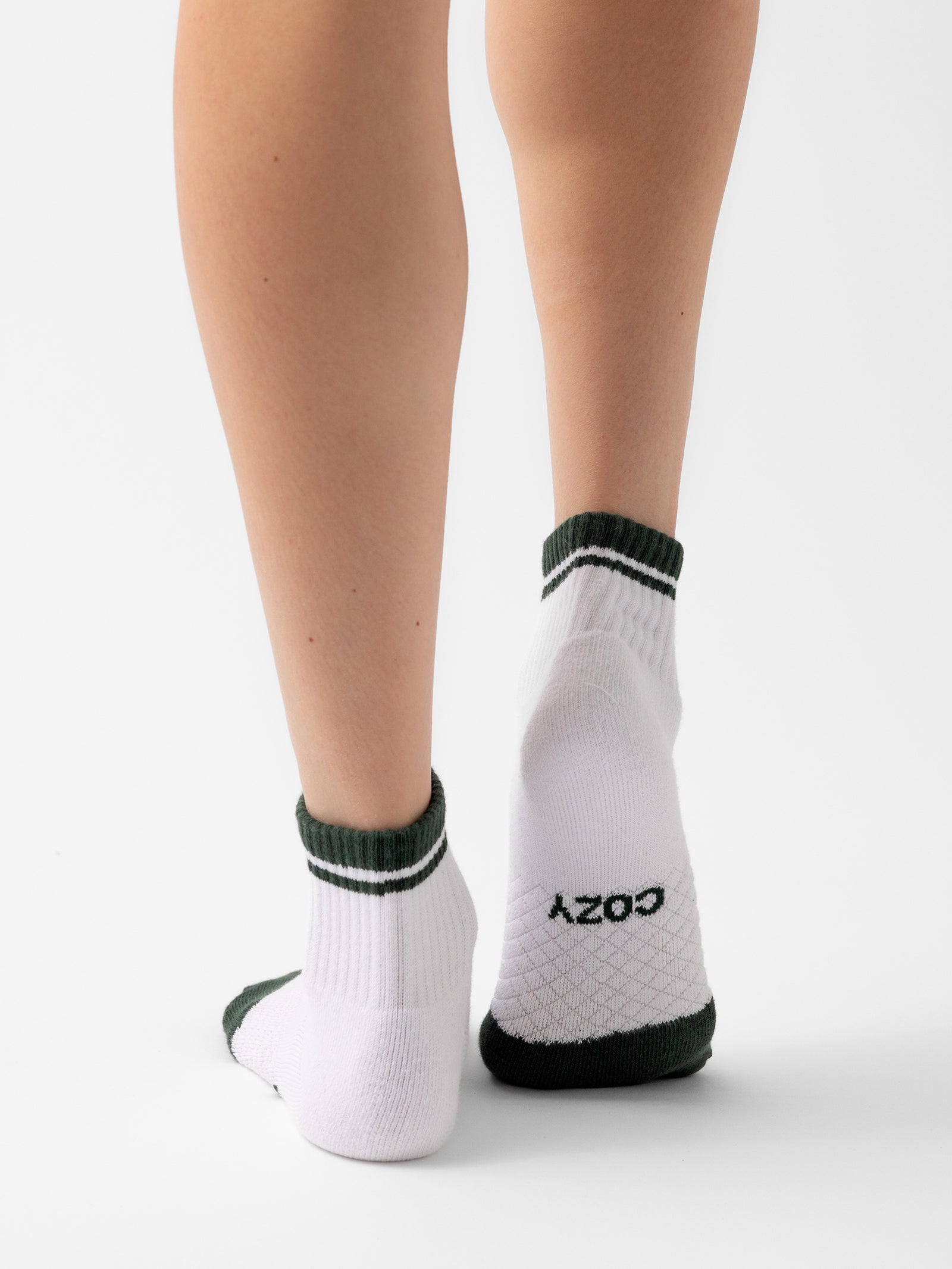 A person stands with their back to the camera, displaying their calves and feet adorned in white socks with green accents from the Cozy Earth Essential Quarter Sock 4-Pack. The word "COZY" is clearly visible on the bottom of the right sock against a plain white background. 