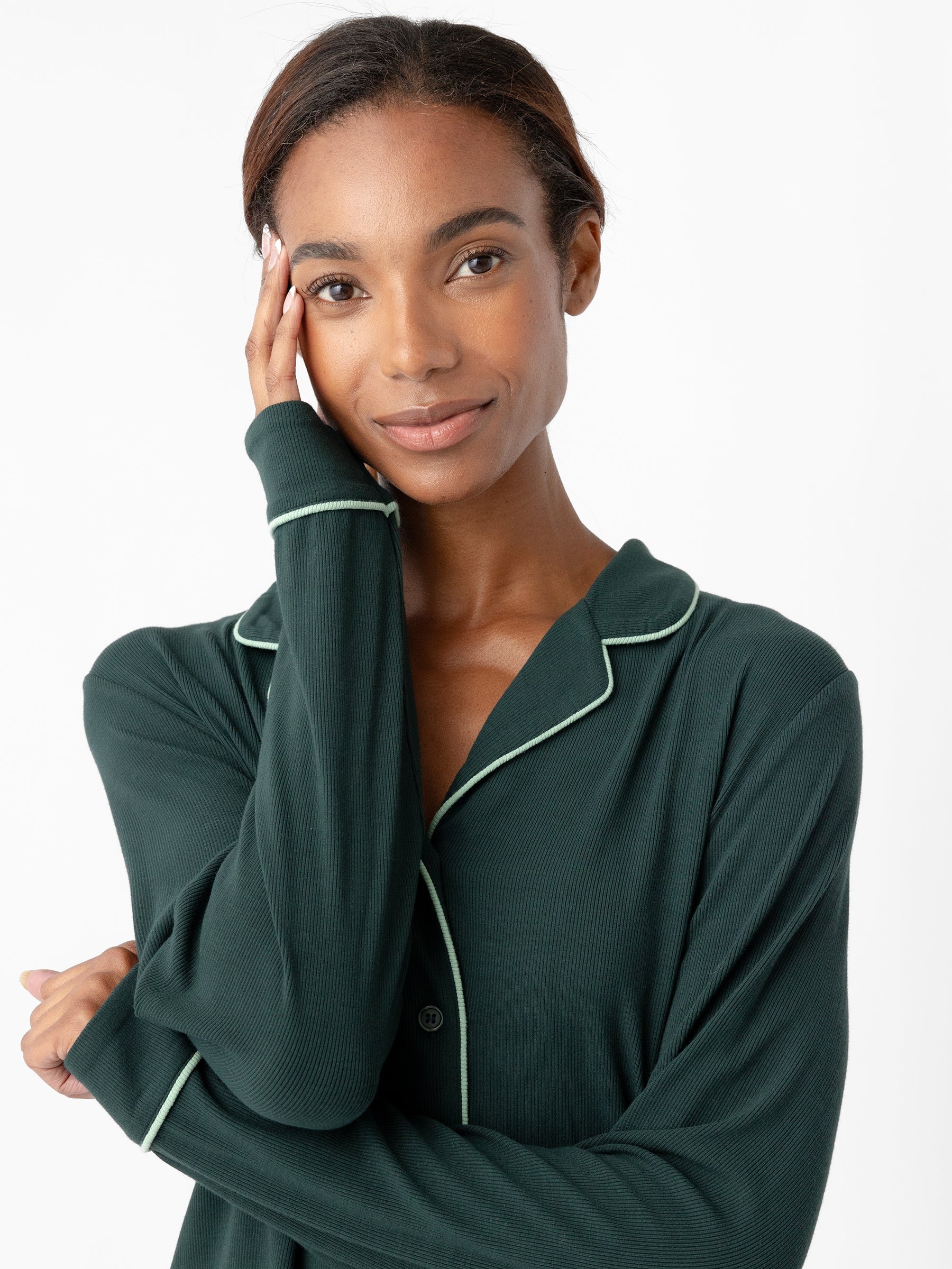A person with dark hair looks at the camera with a gentle smile. They are wearing Cozy Earth's Women's Bamboo Rib Knit Classic Long Sleeve Pajama Top in dark green with light-colored trimming and have one hand touching their cheek. The background is white. 