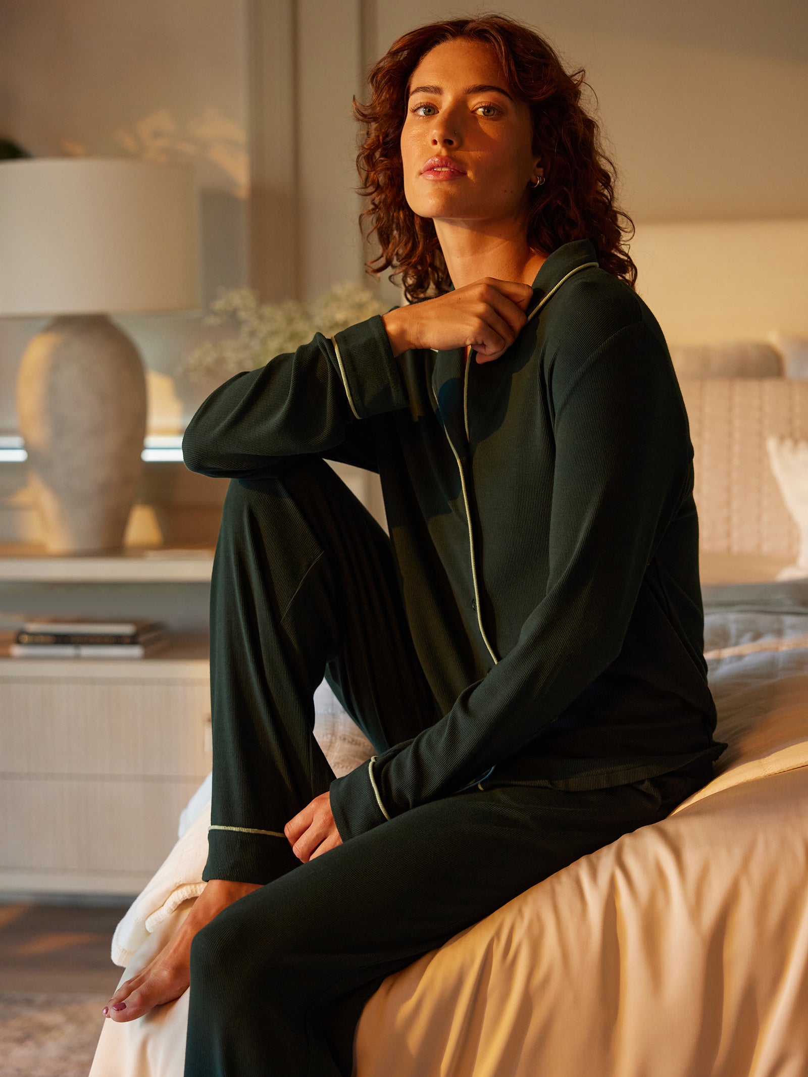 A person with curly hair sits on the edge of a bed, wearing Cozy Earth's Women's Bamboo Rib Knit Classic Pajama Pant in dark green with light piping. They have a thoughtful expression, resting one hand on their knee. The room is warmly lit by a table lamp, with a decorative vase visible in the background. 