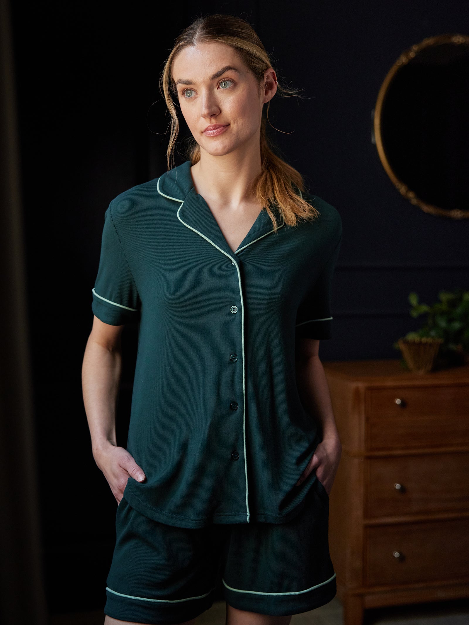 A person with light hair tied back in a ponytail, wearing the Cozy Earth Women's Bamboo Rib Knit Classic Short Sleeve Pajama Top in dark green with white piping, stands with hands in pockets. The backdrop features a wooden dresser, a circular mirror, and a small plant in a pot. 