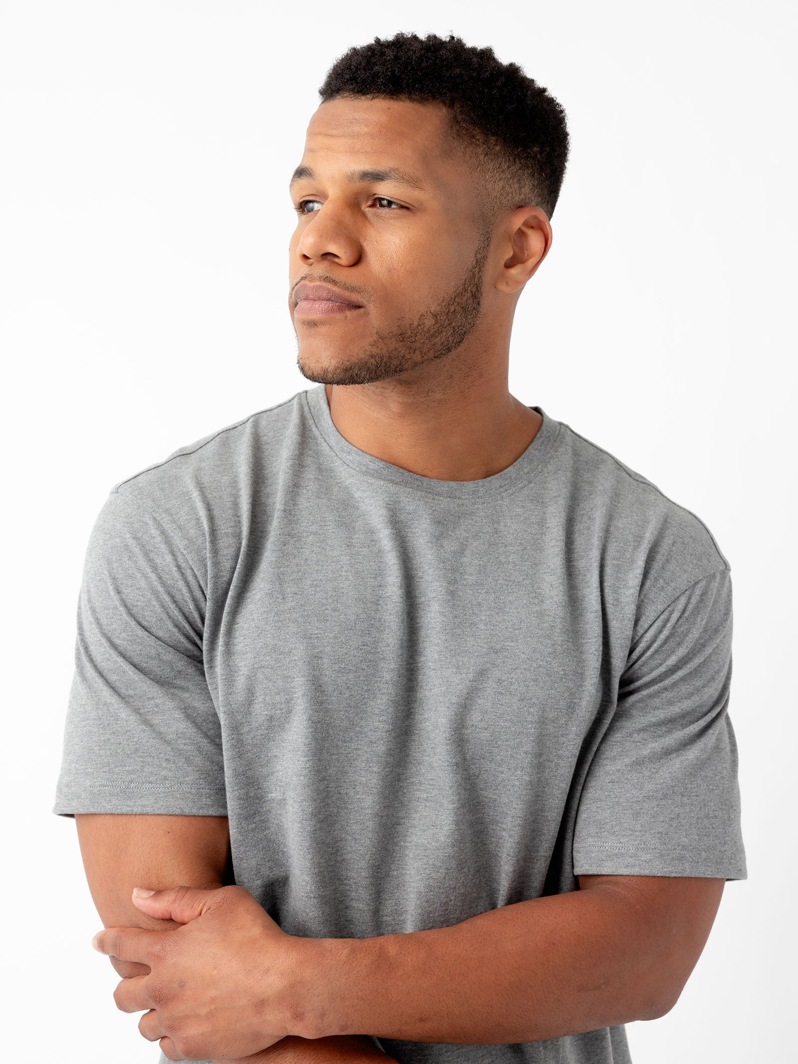 A man with short curly hair and a trimmed beard is standing against a plain white background, wearing the Cozy Earth Men's All Day Tee in gray. He is looking to his left with his arms crossed over his chest. 