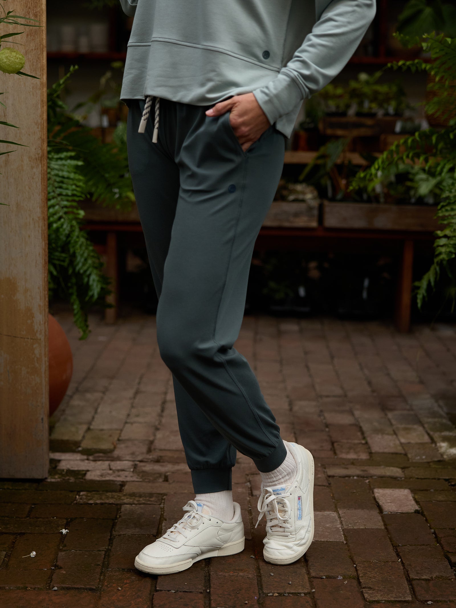 Someone wearing Cozy Earth's Women's Studio Jogger and a matching long-sleeve top, along with white sneakers, stands on a brick path with one leg slightly crossed in front of the other. Lush green plants and wooden furniture can be seen in the background. 