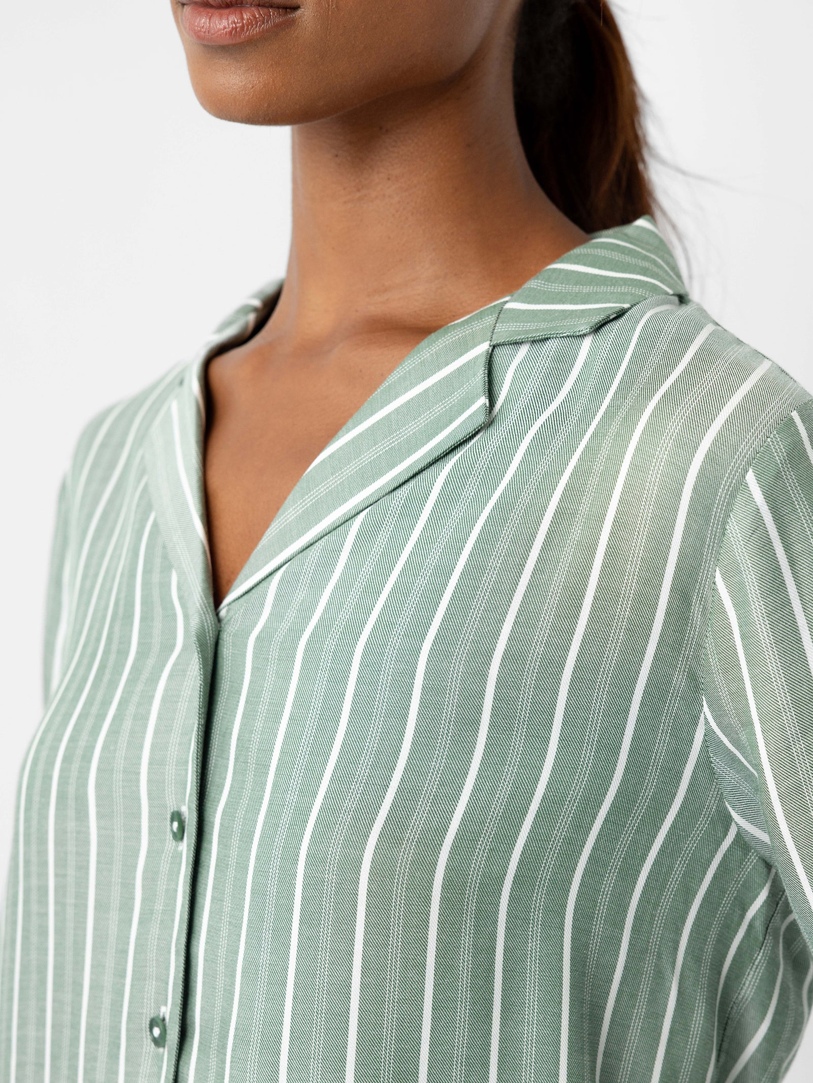 A person wearing Cozy Earth's Women's Soft Woven Long Sleeve Pajama Top is shown. The top features a green and white striped pattern with a collar and a button-down design. The individual has long, dark hair pulled back and appears from the neck to mid-torso against a white background. 