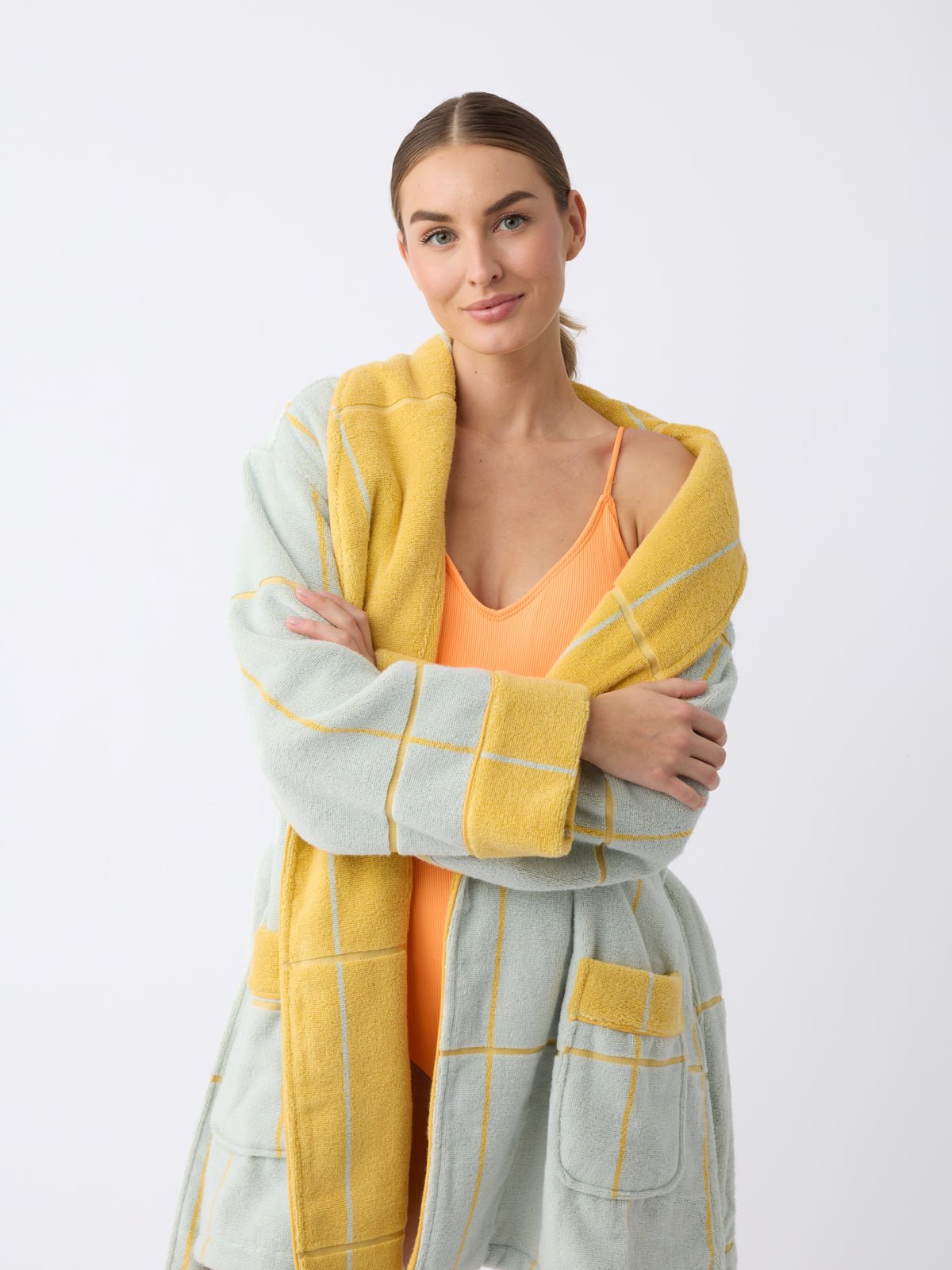 A person with long hair stands against a plain background, smiling with arms crossed while wearing a Cozy Earth Windowpane Resort Robe in light blue with yellow grid patterns and a yellow inner lining over an orange swimsuit. 