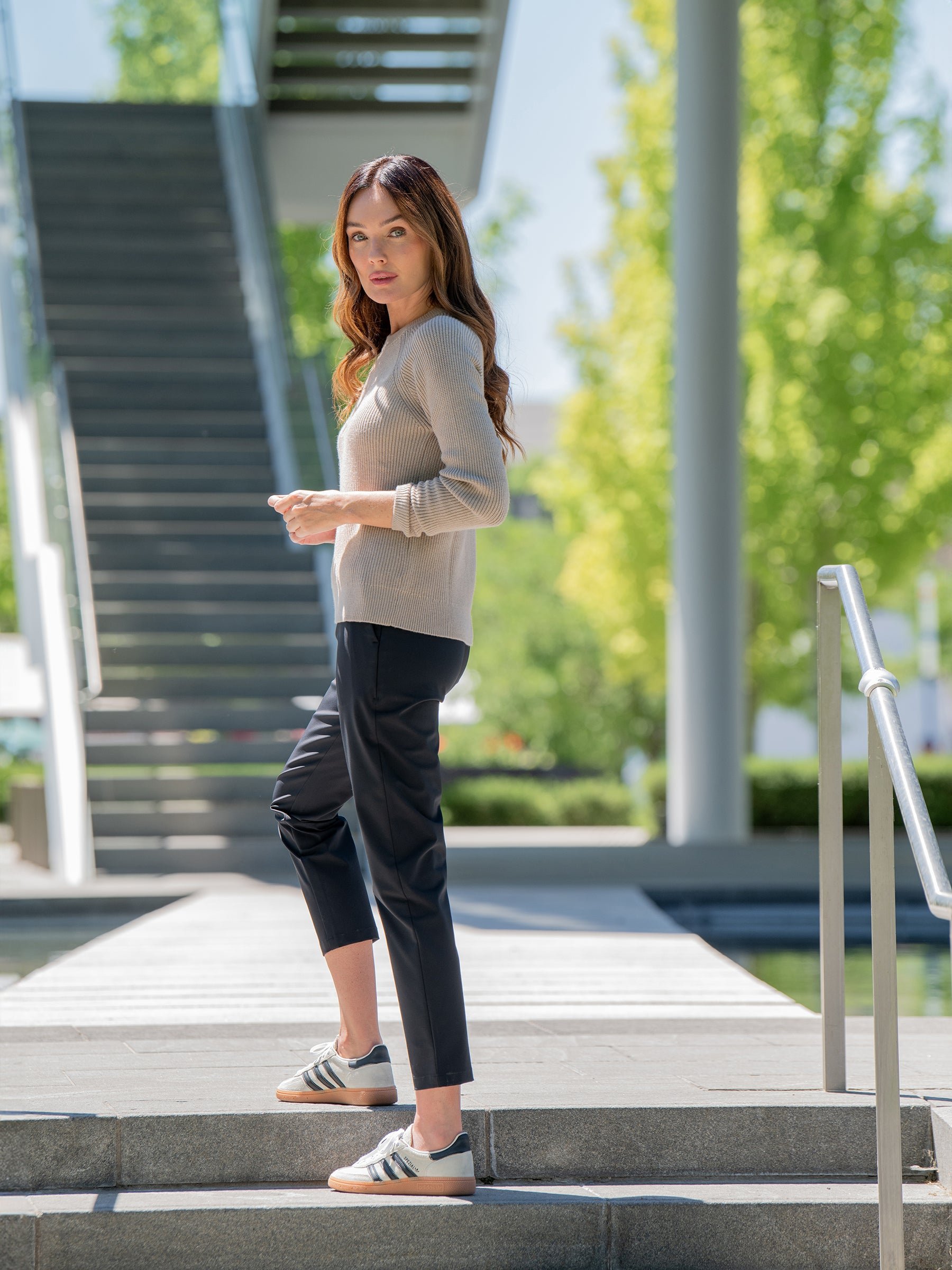 A woman with long brown hair stands on an outdoor staircase, wearing a beige Women's Classic Crewneck by Cozy Earth, black pants, and gray Adidas sneakers. She looks over her shoulder with a serious expression. The background features a modern structure and green trees. 