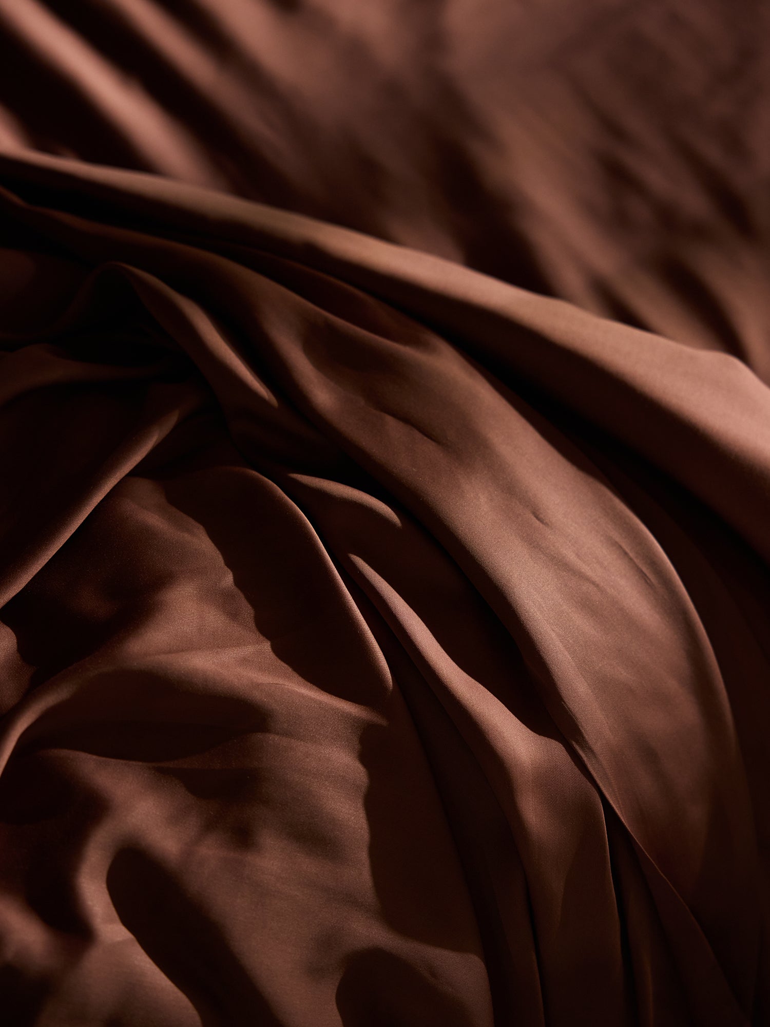 A close-up of the Cozy Earth Bamboo Fitted Sheet reveals its rich, dark brown hue and silky texture. The fabric exhibits slight wrinkles that create smooth folds, with subtle, varying shades of brown as light dances across its surface. 