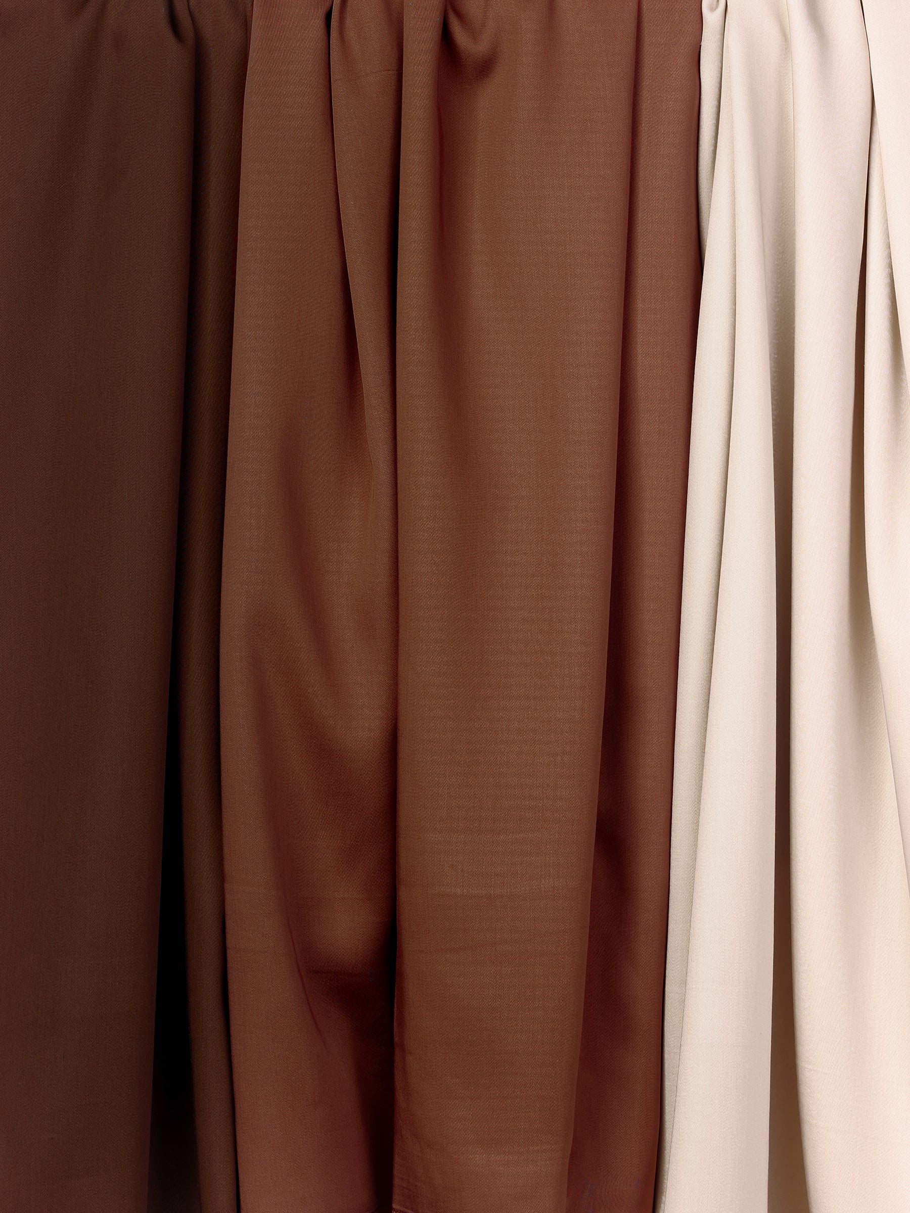 The Bamboo Sheet Set by Cozy Earth features a range of hanging fabrics in various shades of brown and beige. Uniformly draped in vertical folds, each fabric showcases the texture and slight sheen of the material. The colors transition seamlessly from dark brown on the left to a light beige on the right. 