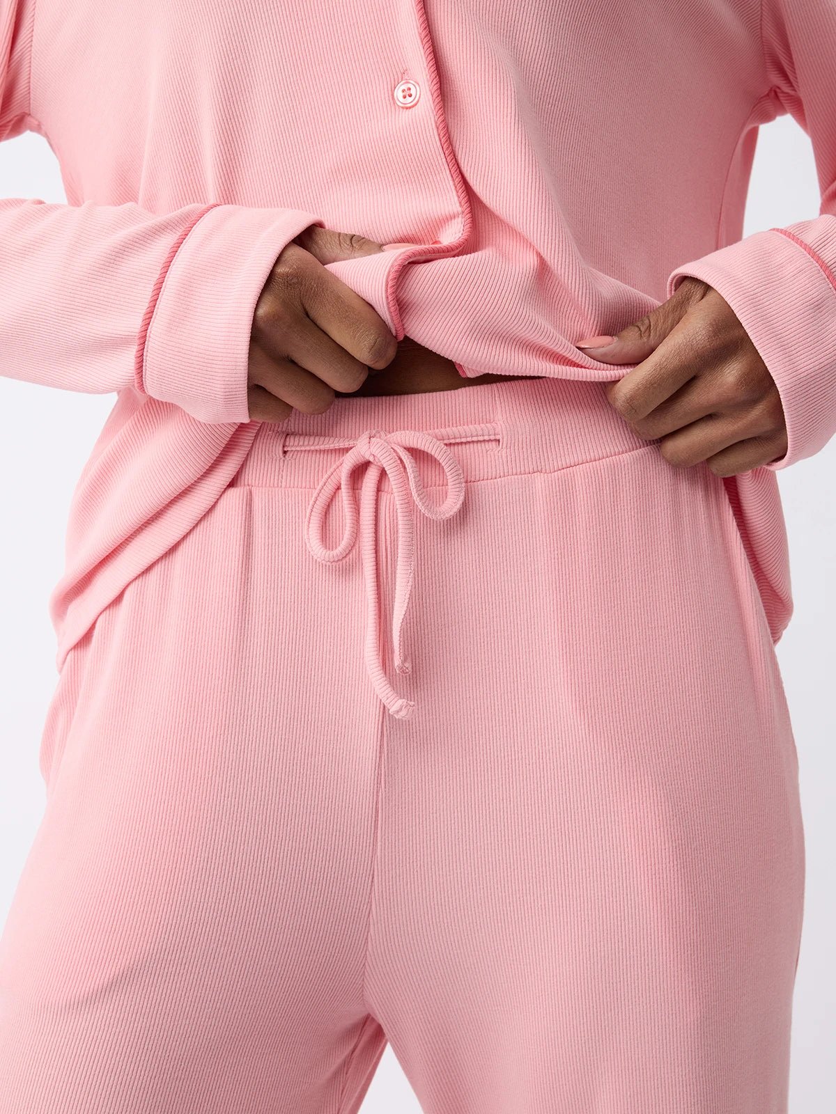 A woman wearing Cozy Earth's Women's Bamboo Rib-Knit Classic Pajama Pant in light pink ribbed fabric, featuring buttons and drawstring pants, slightly pulls the top to showcase its texture. 