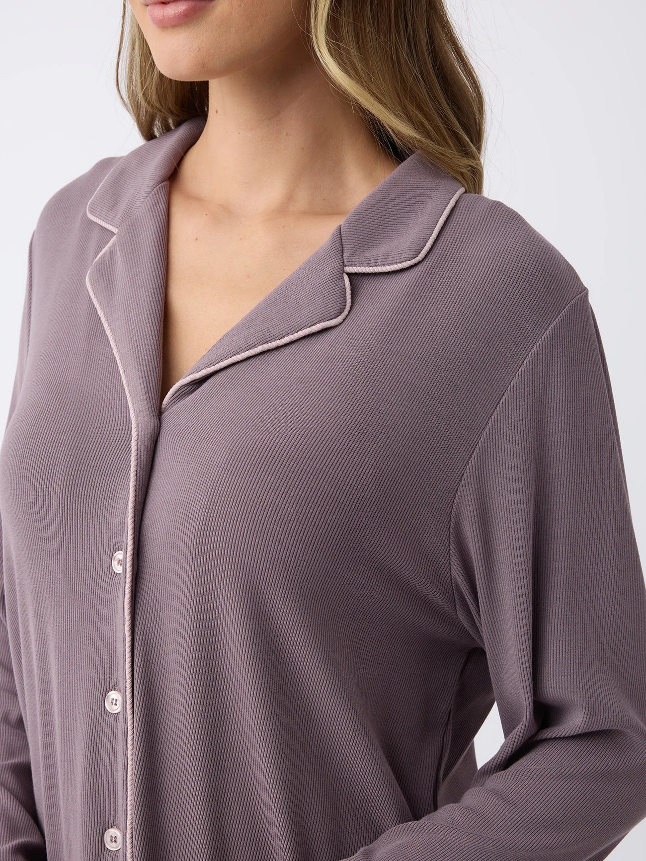 A woman dons a soft, relaxed mauve button-up shirt from Cozy Earth's Rib-Knit Classic Long Sleeve Pajama Set, featuring a notched collar and ribbed texture. Their hair is wavy against a white background. 