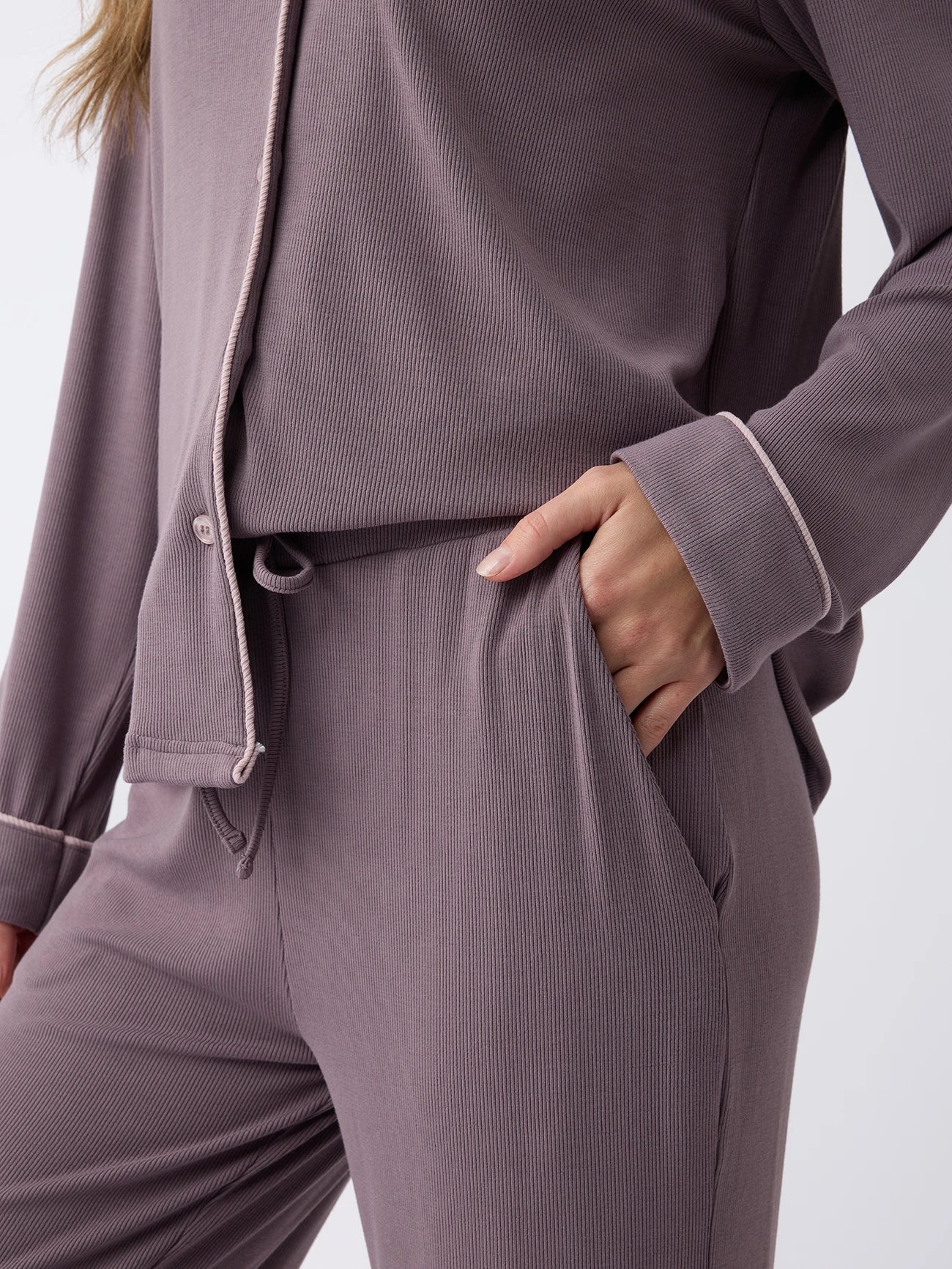 A woman is wearing Cozy Earth's mauve Women's Bamboo Rib-Knit Classic Pajama Pant with light pink piping, one hand in the pocket, and the other arm hanging down against a plain white background. 